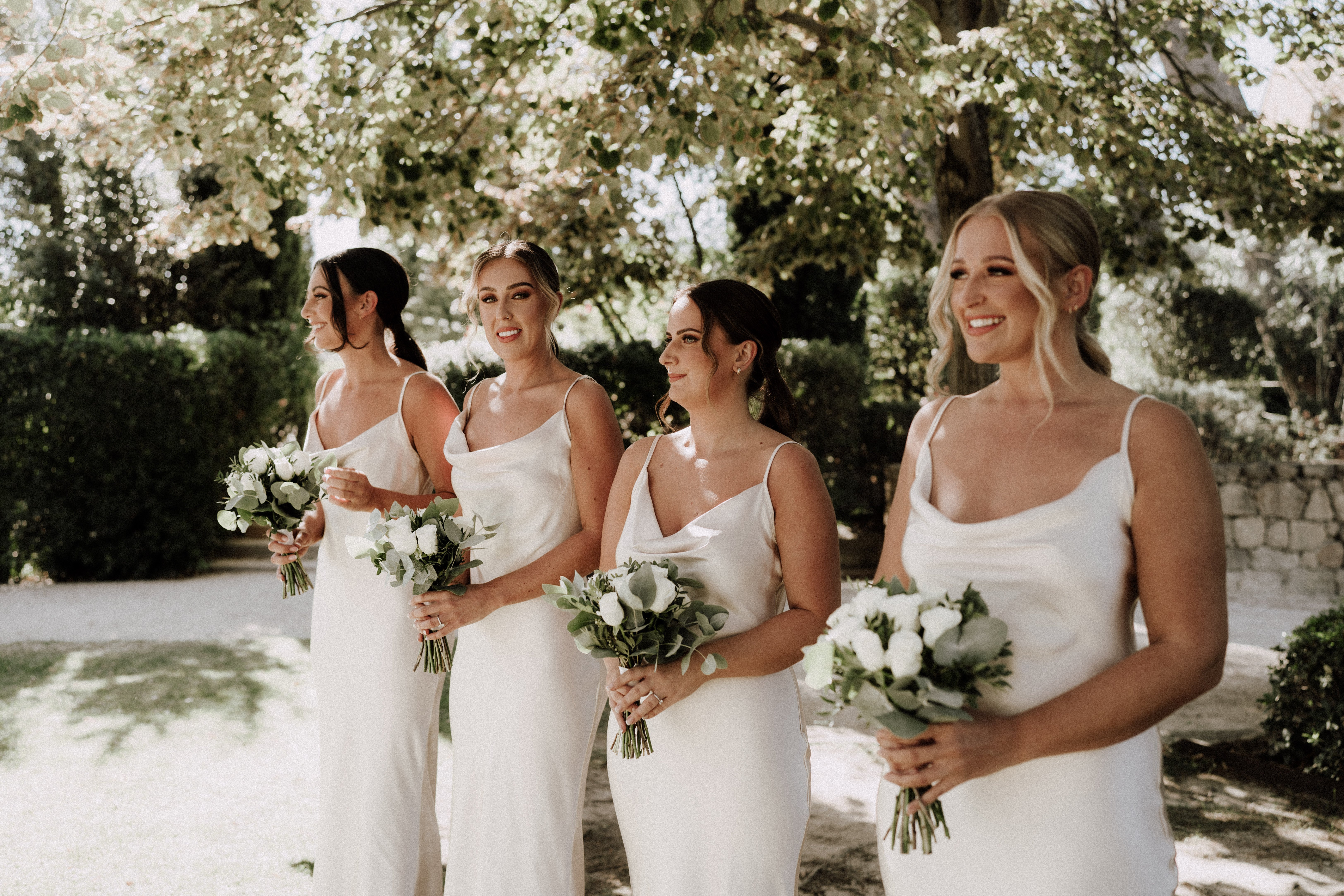 Four bridesmaids stand together outdoors in a garden setting, posed in a loose diagonal line and facing slightly to one side. All four wear matching ivory satin slip dresses with spaghetti straps and a cowl neckline, styled in a sleek, modern minimalist aesthetic. Each holds a small bouquet of white roses and ranunculus with eucalyptus and mixed greenery. The bridesmaids have polished updo and half-up hairstyles, and all wear similar natural glam makeup. The shot is a mid-length portrait taken in bright natural daylight.