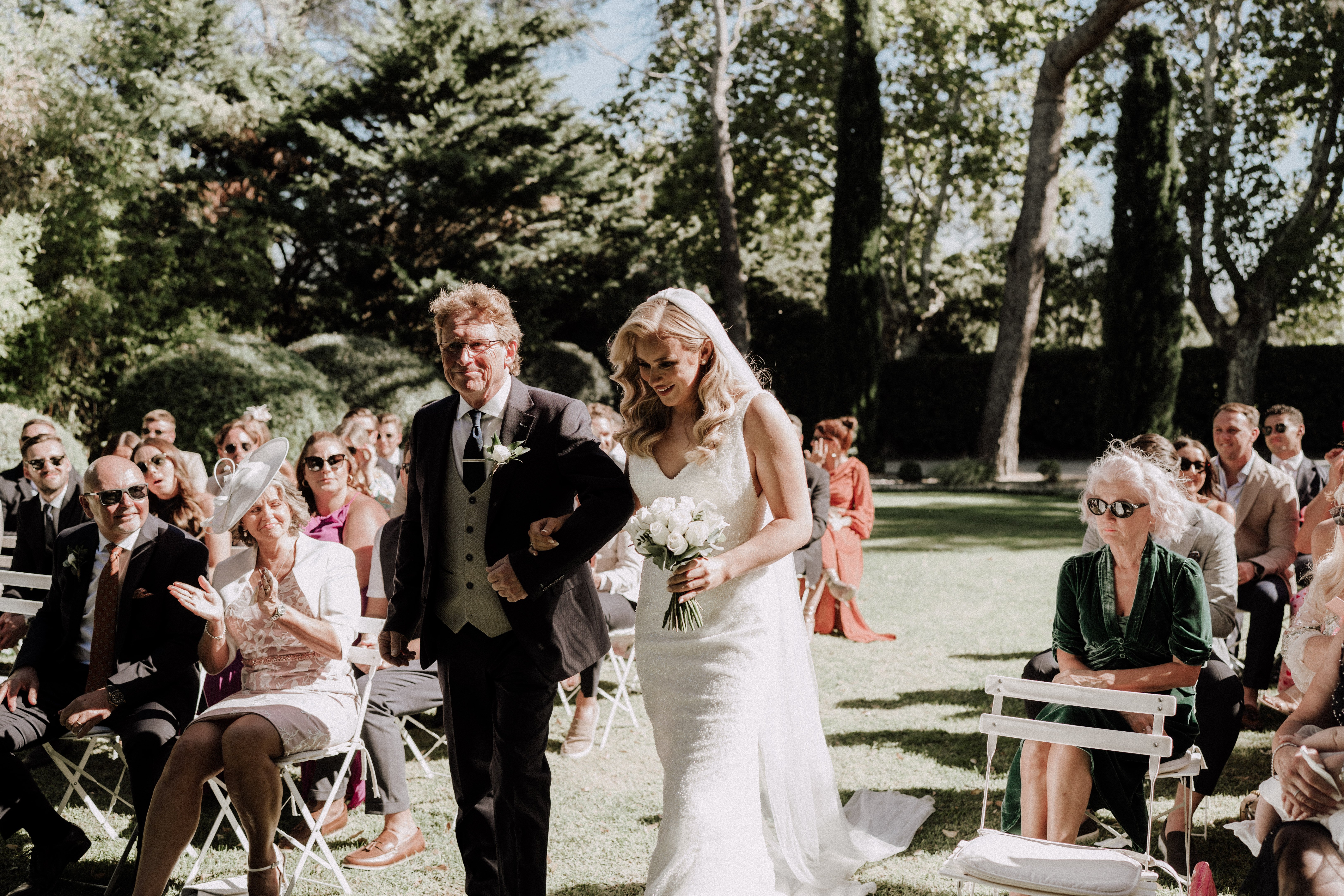 The bride is being walked down the aisle during an outdoor ceremony, escorted by an older man in a dark navy suit with a white boutonnière, likely her father. The bride wears a fitted, sleeveless ivory lace gown with a cathedral-length veil and carries a compact bouquet of white roses with greenery. Guests — approximately 40–50 visible — are seated on white folding chairs arranged in two sections on a lawn, dressed in a range of colorful outfits including a woman in a wide-brim white fascinator hat and a woman in deep green velvet. The setting is a formal garden with tall hedges and mature trees providing a backdrop. The shot is a mid-wide angle taken from ground level at the end of the aisle, capturing the processional moment with the seated guests on both sides reacting warmly.