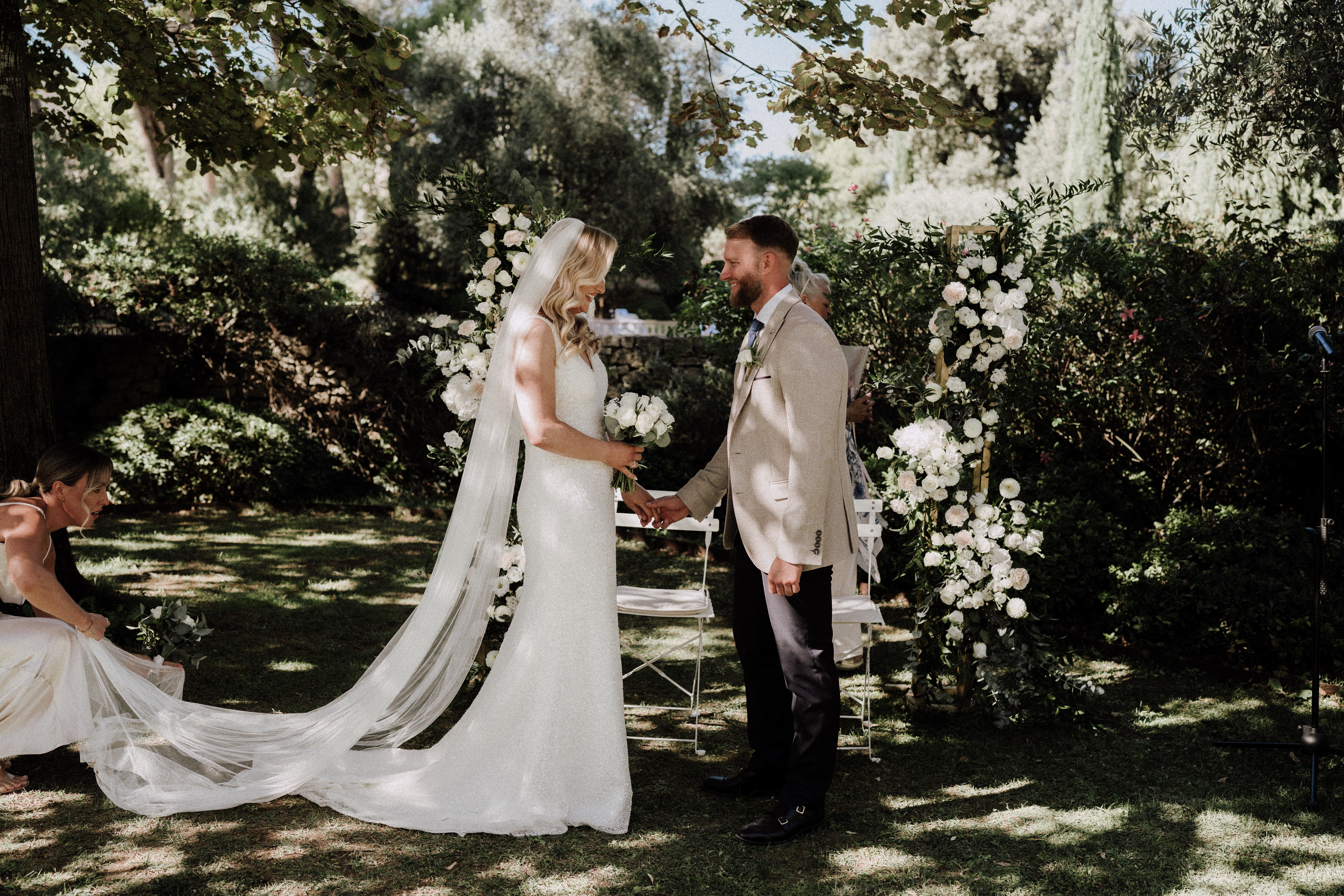 An outdoor wedding ceremony taking place in a lush garden, with the bride and groom holding hands at the altar. The bride wears a fitted ivory sequined gown with a long cathedral-length veil and carries a white and green bouquet of roses and eucalyptus, while the groom wears a sand-colored blazer, dark trousers, and a blue tie. Behind them stands a wooden arch decorated with white roses, peonies, and greenery, flanked by matching floral column arrangements in the same white and green palette. A bridesmaid in a white dress is visible to the left, adjusting the bride's veil train on the grass, and white folding chairs with seated guests are visible in the background. The shot is a medium wide portrait taken in dappled natural sunlight filtering through the trees overhead.