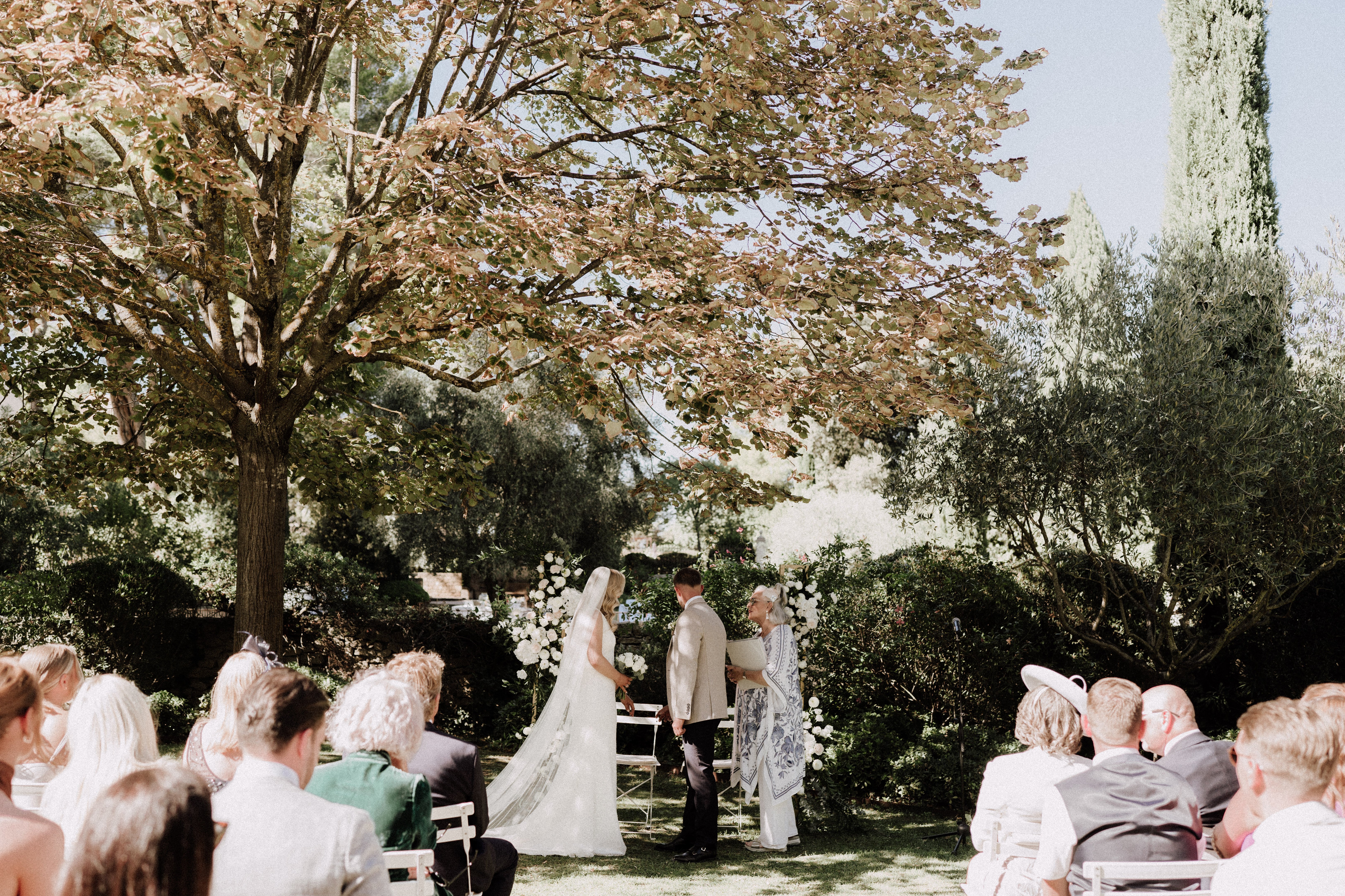 An outdoor wedding ceremony taking place in a garden setting, with the couple standing at the altar facing an officiant dressed in a white outfit with a blue printed shawl. The bride wears a white gown with a long cathedral-length veil, and the groom is dressed in a light beige suit. Two tall floral arrangements of white blooms frame the ceremony space on either side of the couple. Approximately 20–30 guests are seated in white folding chairs arranged in rows on either side of a central aisle, dressed in smart casual to formal attire. The composition is a wide mid-distance shot taken from behind the seated guests, capturing the full ceremony space and surrounding garden landscape including cypress trees in the background.