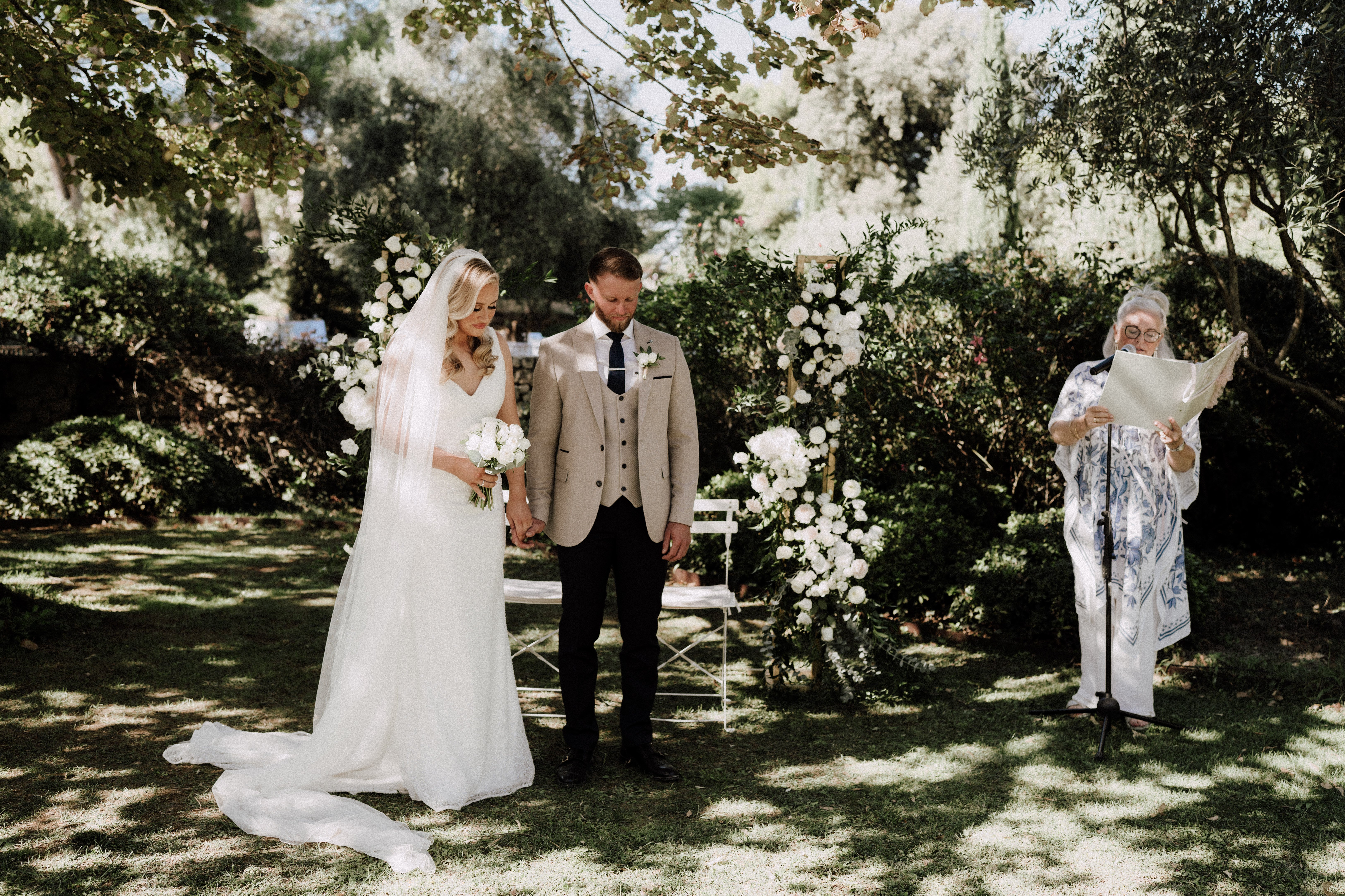 An outdoor garden wedding ceremony showing a bride and groom standing together holding hands, heads bowed, while an officiant reads from papers at a microphone stand to their right. The bride wears a white V-neck fitted gown with a long cathedral-length veil and carries a bouquet of white roses and white peonies; the groom wears a tan three-piece suit with black trousers and a navy tie with a white boutonniere. Behind the couple stands a wooden arch decorated with clusters of white roses and white peonies with greenery, flanked by two additional floral arrangements in the same white palette at ground level. White folding chairs are visible near the arch. The officiant wears a blue and white patterned top. The setting is an intimate garden with dense surrounding hedges and trees providing natural shade. Wide shot composition capturing all three figures and the full ceremony backdrop.