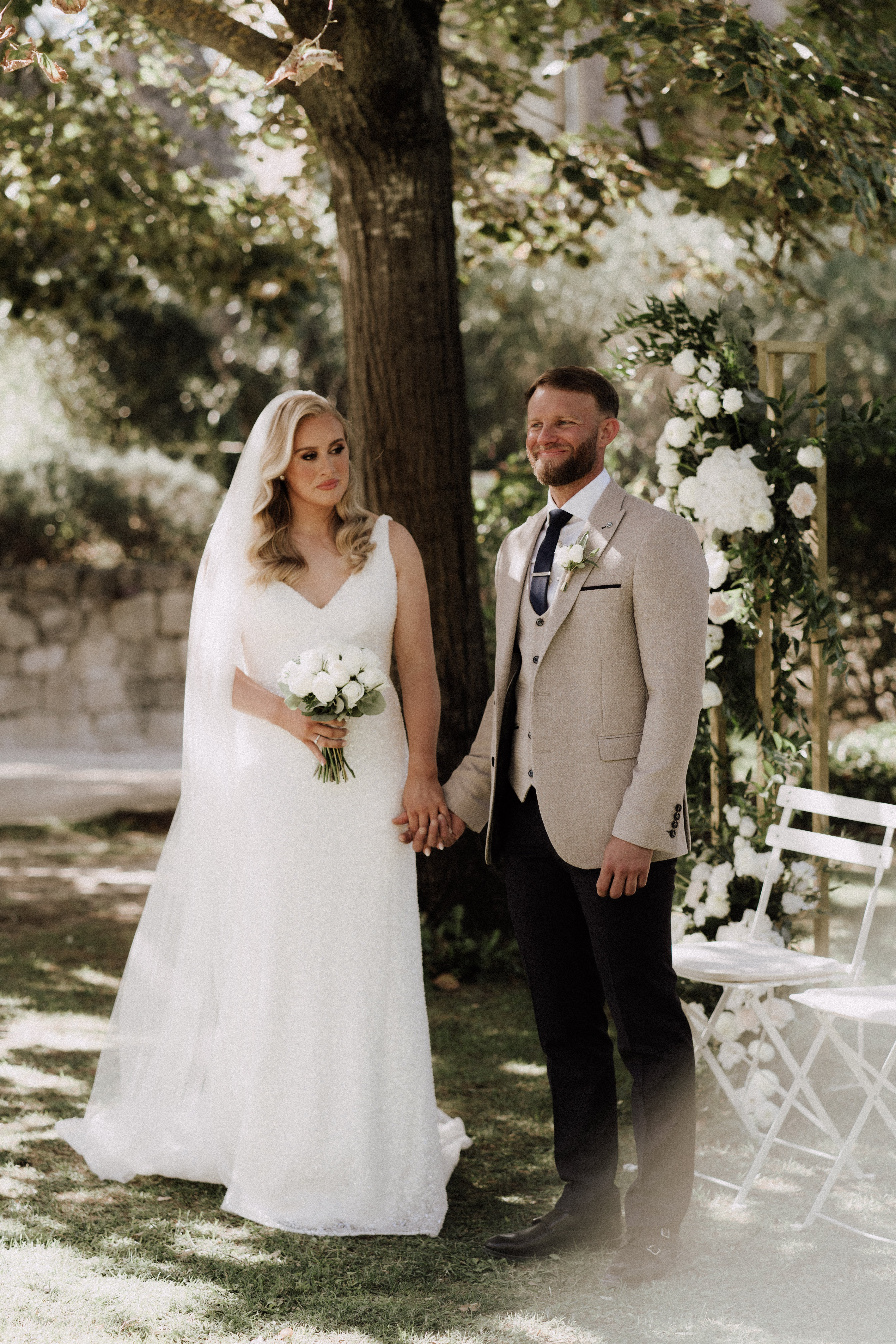 A couple portrait taken outdoors in a garden setting, with the bride and groom standing hand-in-hand beneath a large tree. The bride wears a white V-neck A-line lace gown with a cathedral-length veil and carries a tight round bouquet of white roses and green foliage. The groom wears a sand/beige three-piece suit with dark navy trousers, a white dress shirt, and a navy tie, with a white rose boutonniere. Behind them to the right is a wooden arch decorated with lush greenery and white hydrangeas and roses, alongside white folding chairs indicating an outdoor ceremony setting. The overall decor palette is white and green with a classic, clean aesthetic. Medium portrait shot with a slightly shallow depth of field.