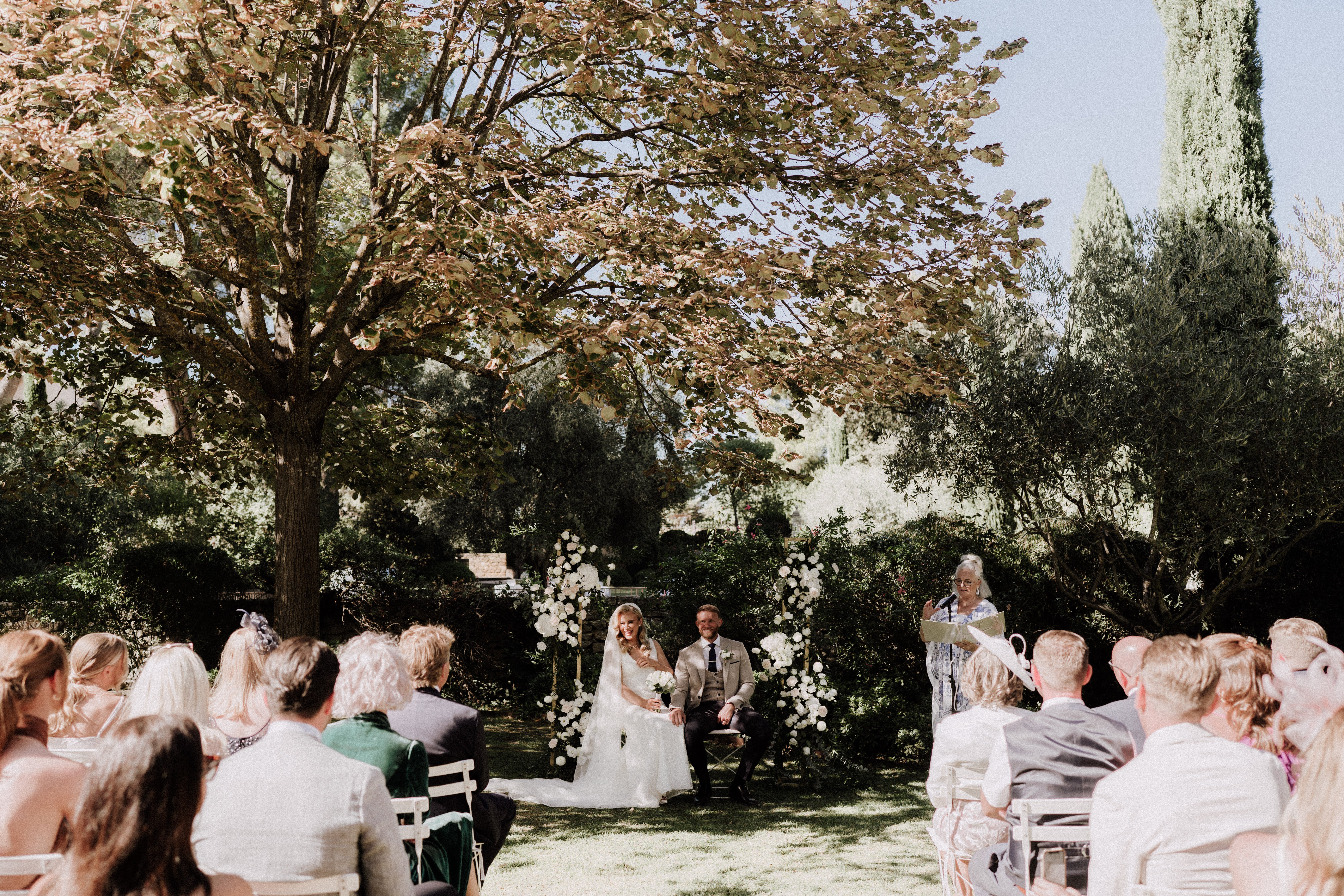 An outdoor wedding ceremony taking place in a garden setting, with the couple seated in front of a gold arch decorated with clusters of white florals — appearing to be white roses and greenery — flanking both sides. The bride wears a white full-length gown and the groom wears a light beige suit with a dark tie; both are smiling and appear to be listening to a reading. A female officiant stands to the right reading from papers at a microphone. Approximately 30–40 guests are seated on white folding chairs arranged in rows on either side of a central aisle, with their backs to the camera in a wide shot. The decor palette is white and gold with a classic garden style.