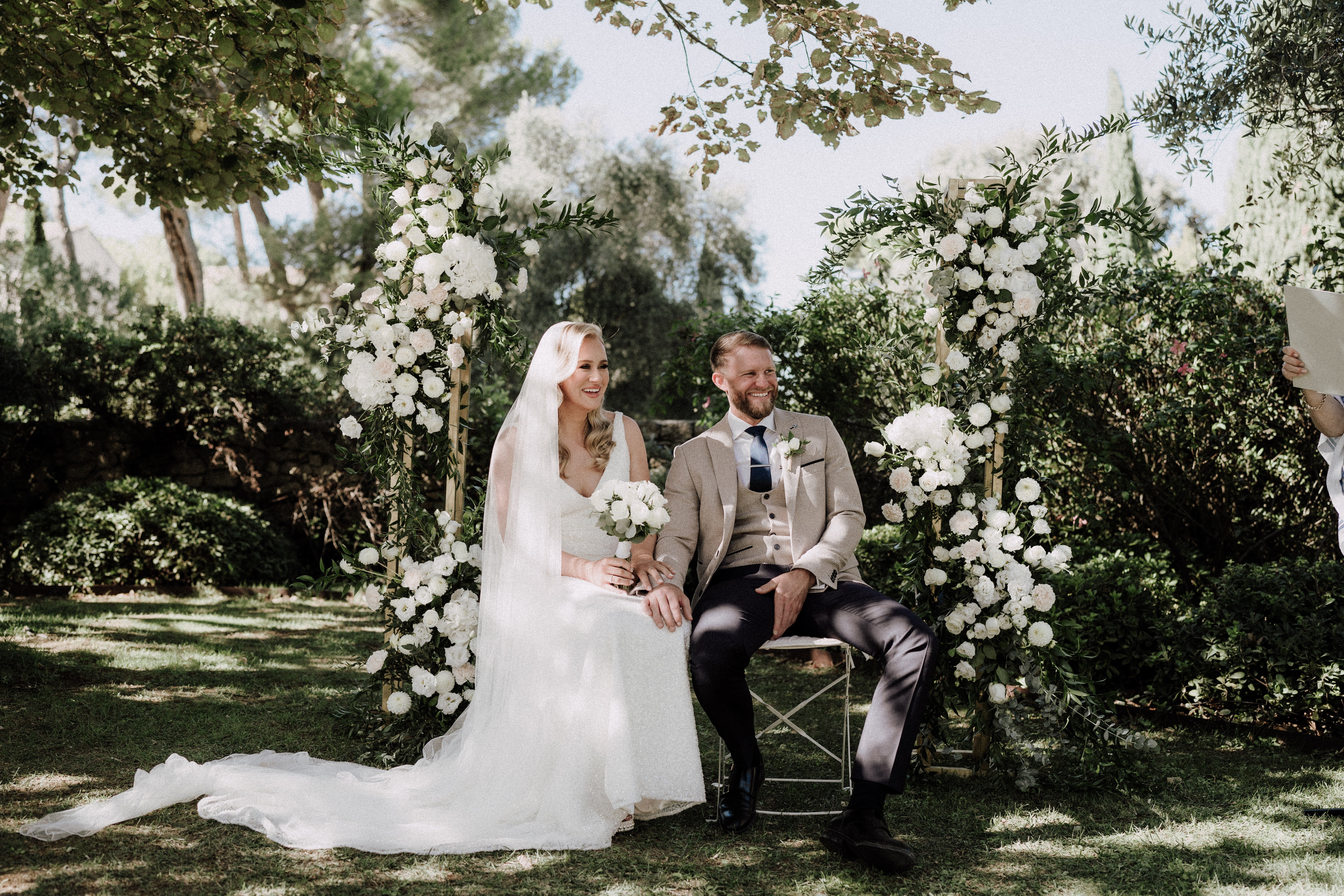 The bride and groom are seated during an outdoor ceremony, laughing and holding hands as an officiant reads to their right. The bride wears a sleeveless white lace gown with a long cathedral-length train and a sheer veil, holding a bouquet of white peonies and soft greenery. The groom is dressed in a tan/beige suit with a matching waistcoat, dark trousers, and a navy blue tie with a white boutonniere. They are flanked by two tall wooden arch structures densely decorated with white peonies, white dahlias, and lush green foliage, creating a classic all-white floral palette against the garden setting. The shot is a medium-wide portrait taken at eye level in a manicured garden with trimmed hedges and mature trees.