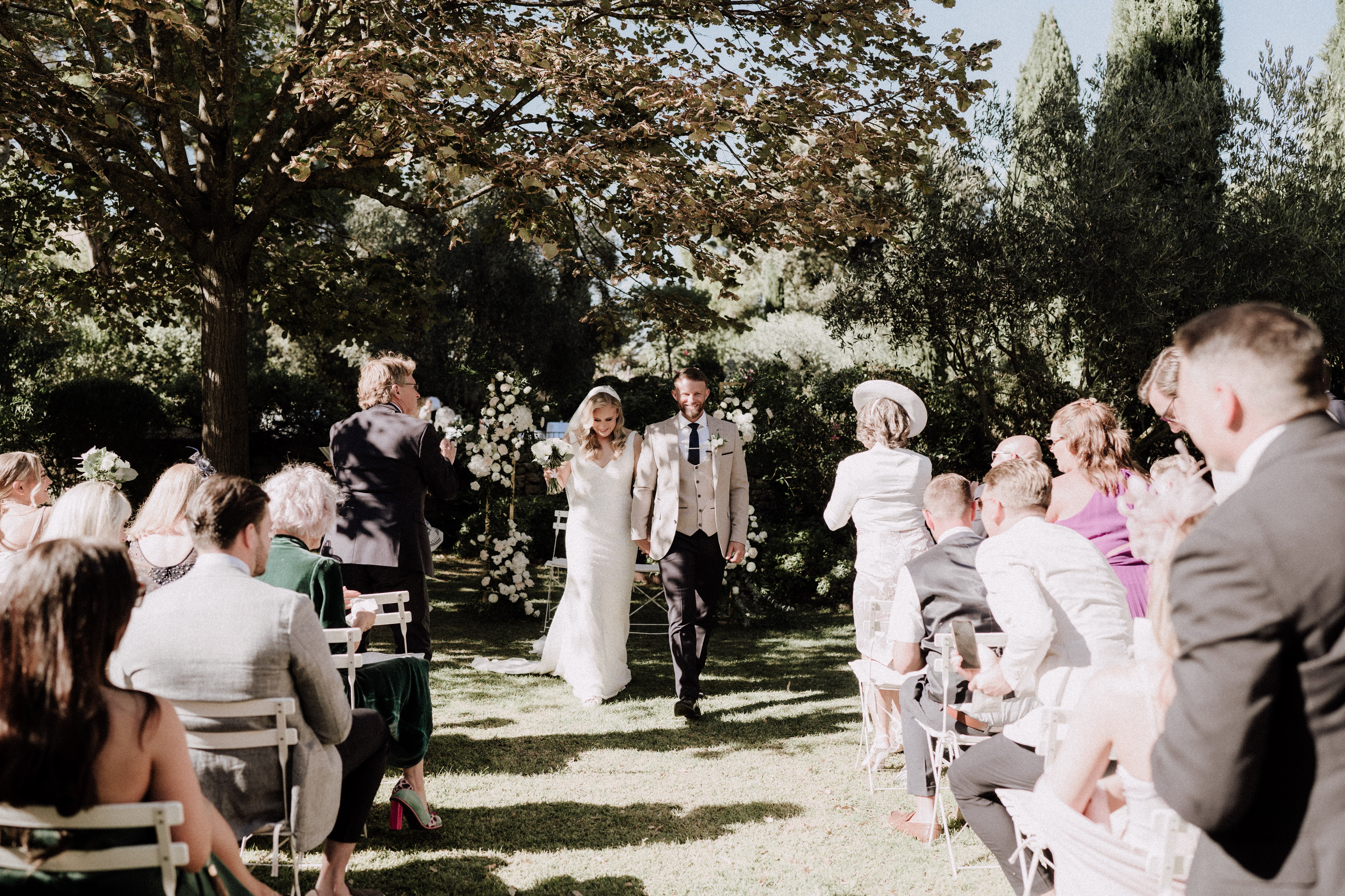 The newlywed couple walks back up the aisle together following an outdoor garden ceremony, with approximately 30 guests seated on white folding chairs on either side. The bride wears a fitted ivory sleeveless gown and holds a small bouquet of white flowers, while the groom wears a light tan/champagne suit jacket with dark navy trousers and a navy tie with a white buttonhole. In the background, a gold arch decorated with white floral arrangements — appearing to include white roses and greenery — marks the ceremony altar. Guests are dressed in a mix of smart-casual outfits including a woman in a magenta dress and another in a wide-brimmed cream hat. The shot is a wide mid-distance perspective taken from behind the seated guests, looking down the aisle toward the couple.