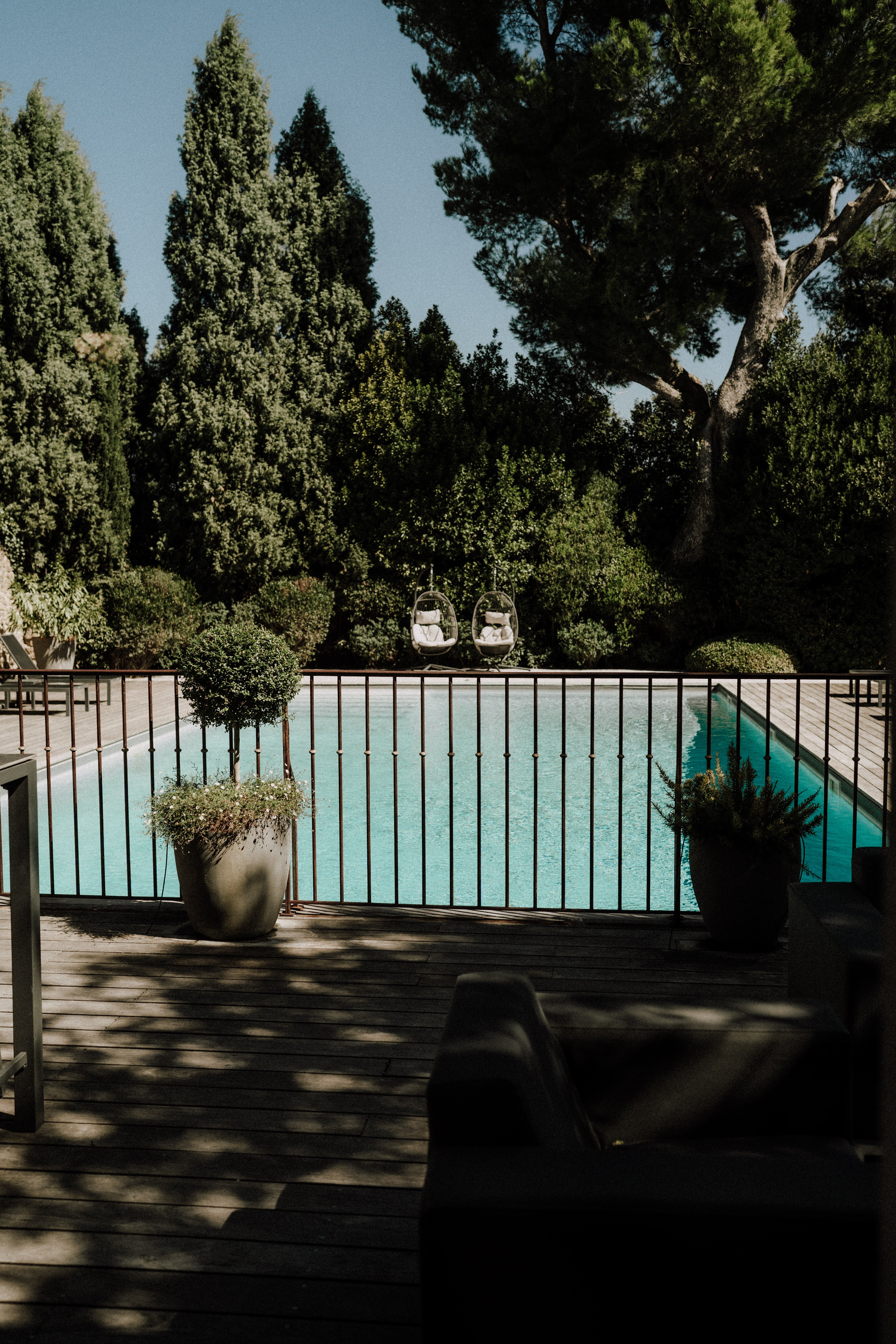 A wide shot of an outdoor pool area at a French property venue, with no people visible. The scene features a rectangular pool with turquoise water, enclosed by a dark wrought-iron railing, viewed from a wooden deck terrace in the foreground. Large cream ceramic planters hold flowering plants and a clipped topiary ball on either side of the deck. Two hanging egg-style chairs with white cushions are positioned on the far side of the pool. The surrounding garden includes tall cypress trees and mature pine trees. Potential venue feature image.