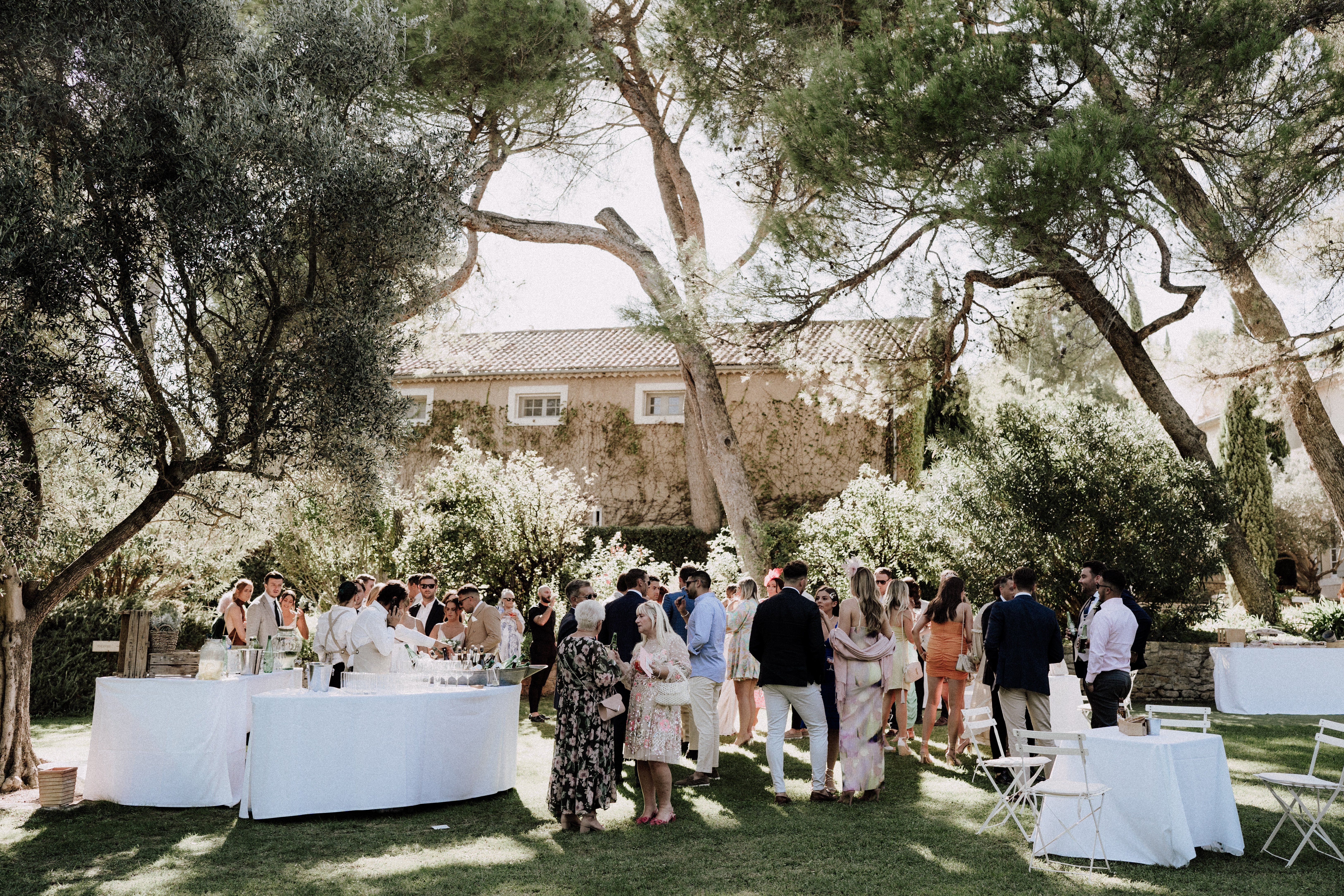 An outdoor cocktail hour taking place on a lawn in front of a traditional Provençal mas or farmhouse with terracotta roof tiles and ivy-covered stone walls. Approximately 40–50 guests mingle in small groups across the garden, dressed in summer wedding attire including floral maxi dresses, linen suits, and cocktail dresses in a mix of navy, orange, white, and floral prints. Two round tables draped in white linen are visible on the left, set with champagne flutes and what appears to be a drinks service station with ice buckets. White folding bistro chairs are arranged to the right side. The scene is lit by bright midday sun filtering through mature olive and pine trees, creating dappled light across the lawn. Wide shot capturing the full scene and venue grounds in the background.