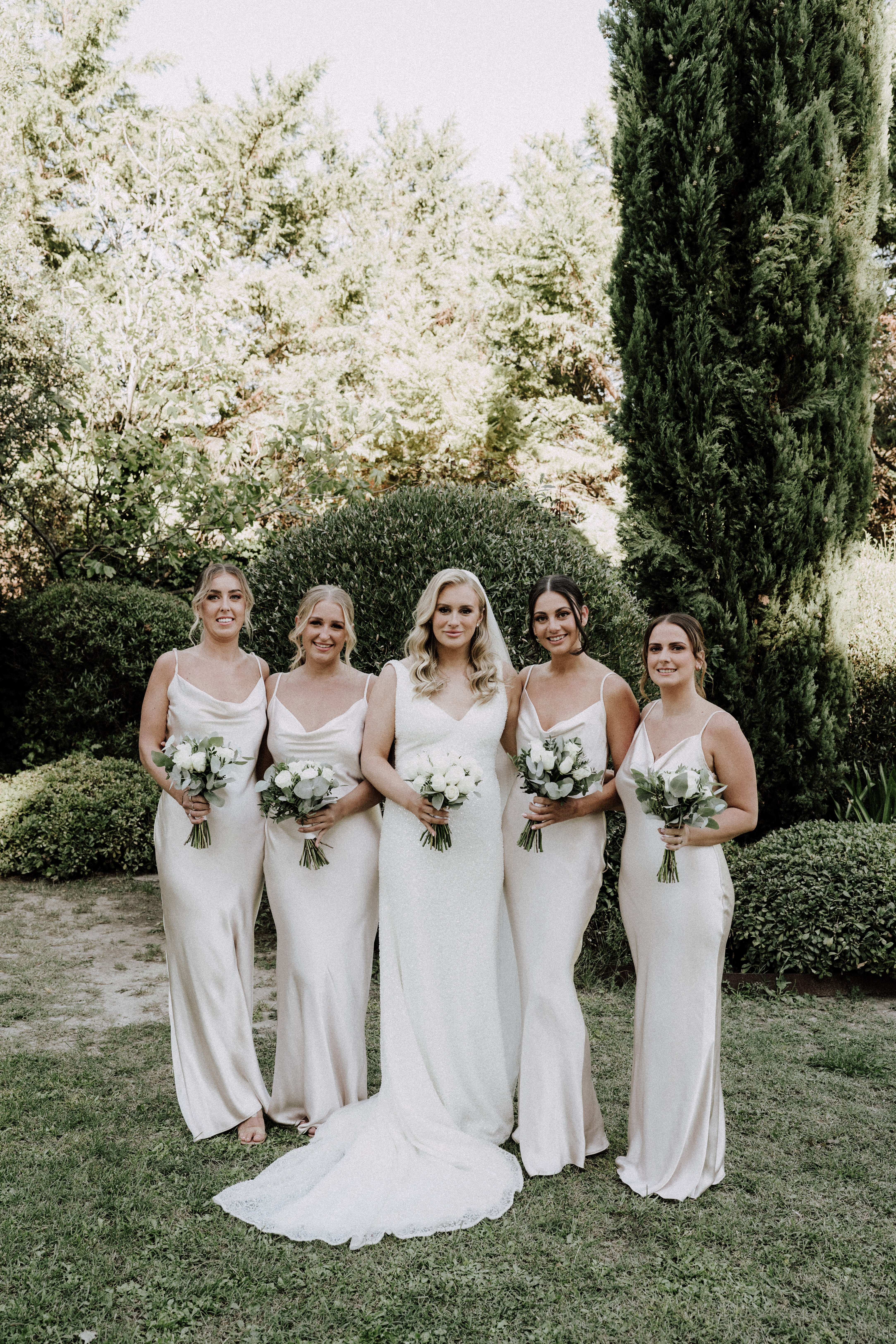 A bridal party portrait taken outdoors in a manicured garden setting with tall cypress trees and trimmed hedges in the background. The bride stands in the center wearing a fitted white lace gown with a small train and V-neckline, flanked by four bridesmaids in champagne-toned satin slip dresses with cowl necklines and thin spaghetti straps. All five women hold bouquets of white roses and ranunculus with eucalyptus greenery. The overall styling palette is white and champagne with a modern, minimalist aesthetic, and the image is a medium full-length group portrait shot in natural daylight.