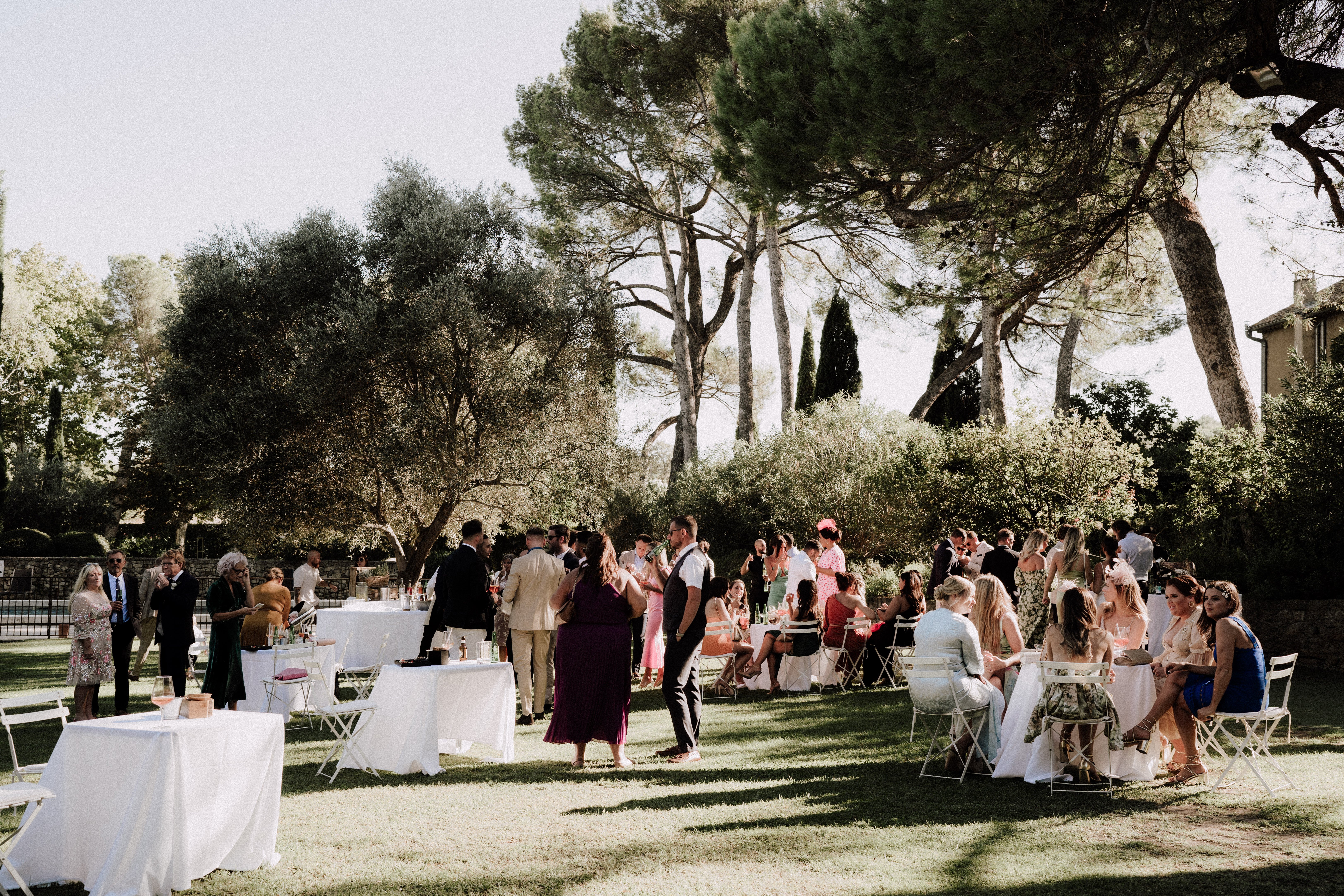 An outdoor cocktail hour taking place on a lawn at what appears to be a French estate or domaine, with approximately 40-50 guests mingling and seated. The setting features white cocktail tables with white linen cloths, white folding chairs, and small bistro-style tables arranged across a grassy area. Guests are dressed in a mix of summer attire including a magenta maxi dress, cobalt blue dress, floral prints, and neutral linen suits, with one guest wearing a notable pink fascinator. A stone building is partially visible to the right edge of the frame. The wide shot captures the full scene in bright midday sunlight, with dappled shade cast across the lawn.