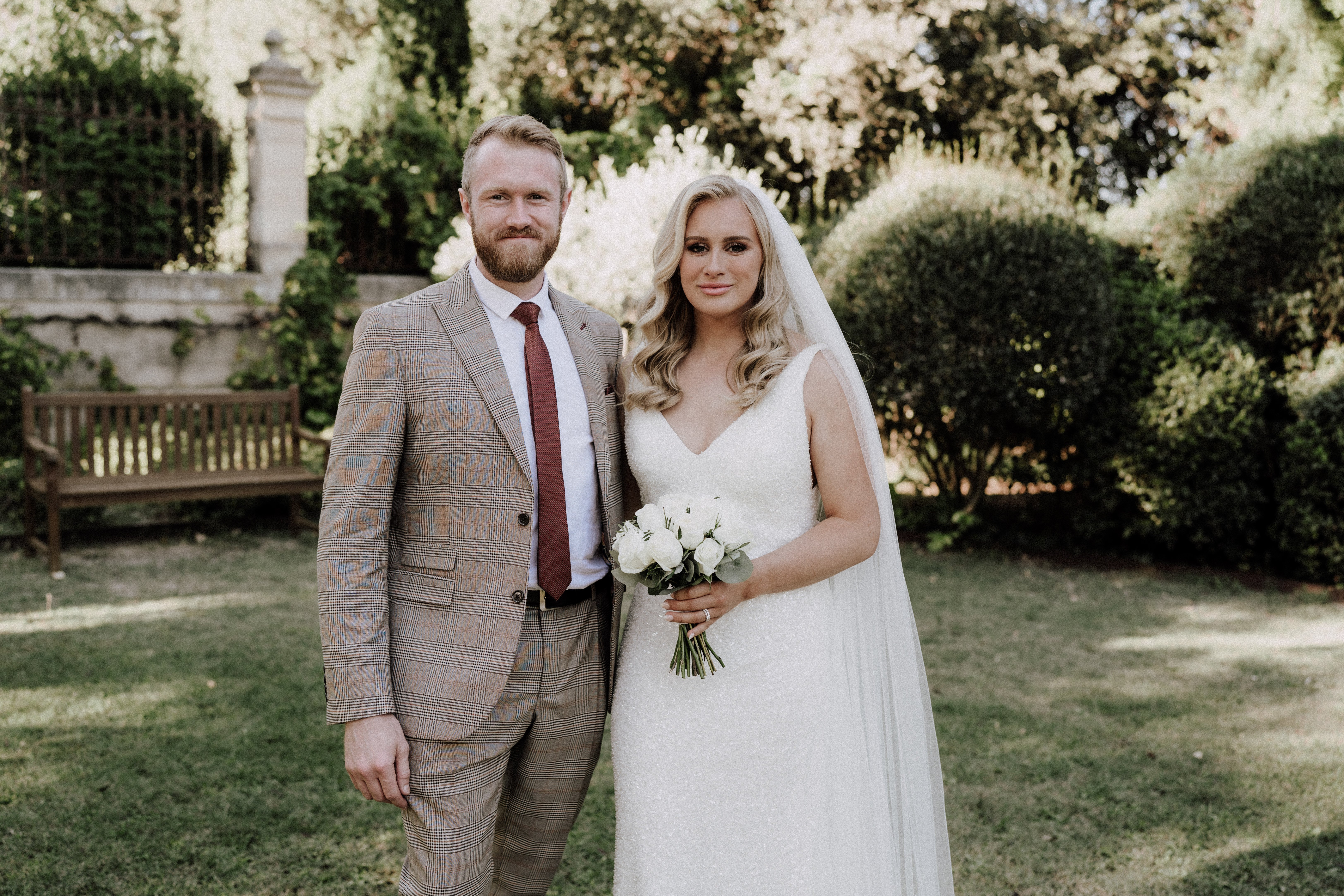 A couple portrait taken outdoors in a formal garden setting, with clipped hedges, a wooden bench, and a stone pillar visible in the background. The bride wears a fitted ivory beaded gown with a V-neckline and a long sheer veil, and carries a compact bouquet of white roses with greenery. The groom wears a tan glen plaid suit with a burgundy tie and white dress shirt. The composition is a mid-length portrait with both subjects facing the camera directly.