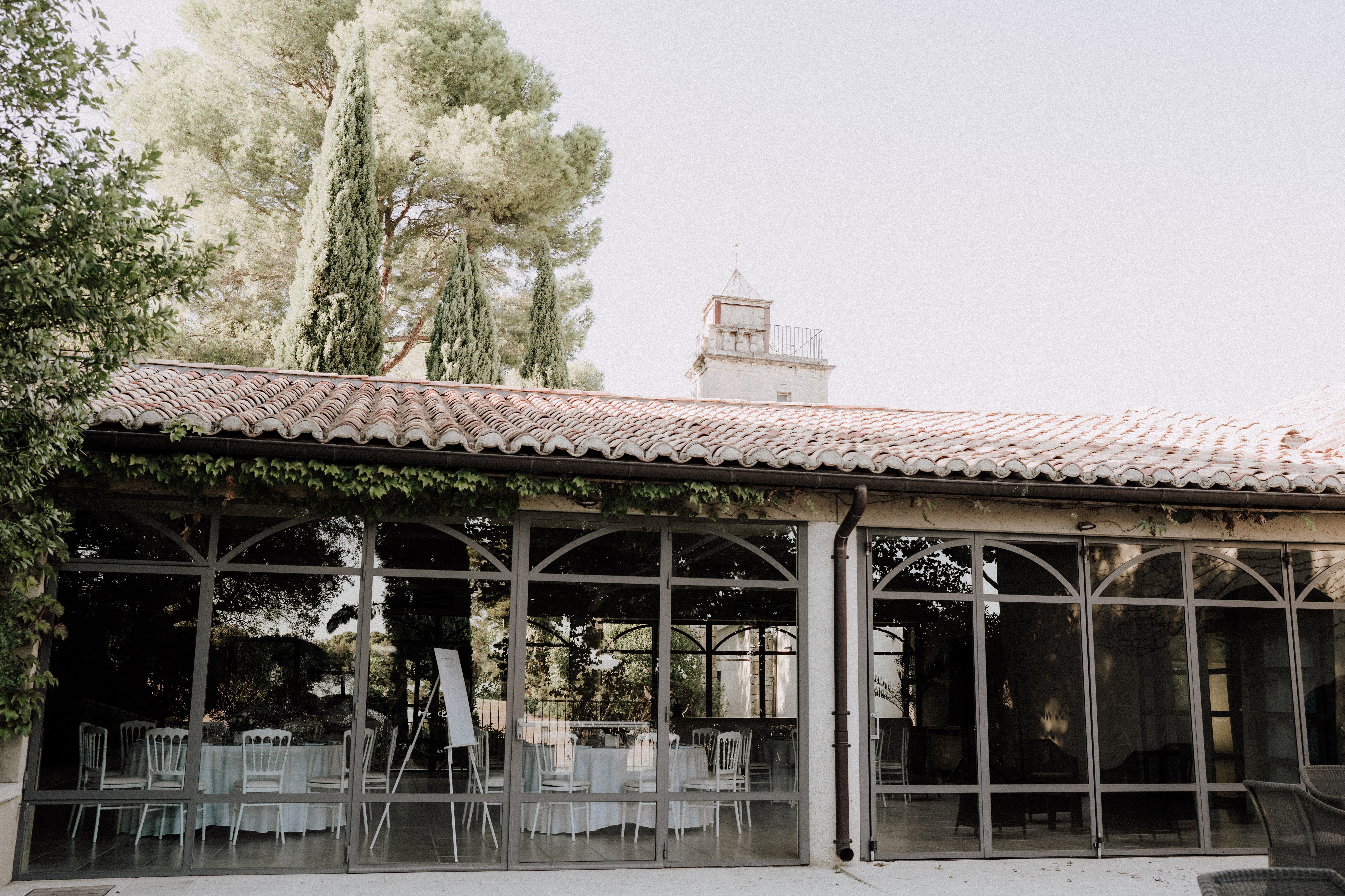 A wide exterior shot of a glass-walled reception pavilion at a French property, viewed from the terrace outside. The structure features terracotta roof tiles, arched metal-framed glass panels, and climbing ivy along the roofline, consistent with a classic Provençal mas or domaine style. Inside the pavilion, round tables are dressed in white linens and surrounded by white Napoléon chairs, with a seating chart displayed on an easel — indicating the space is set up and ready for a wedding reception. A tower or belvedere from the main estate building is visible in the background above the roofline. Potential venue feature image.