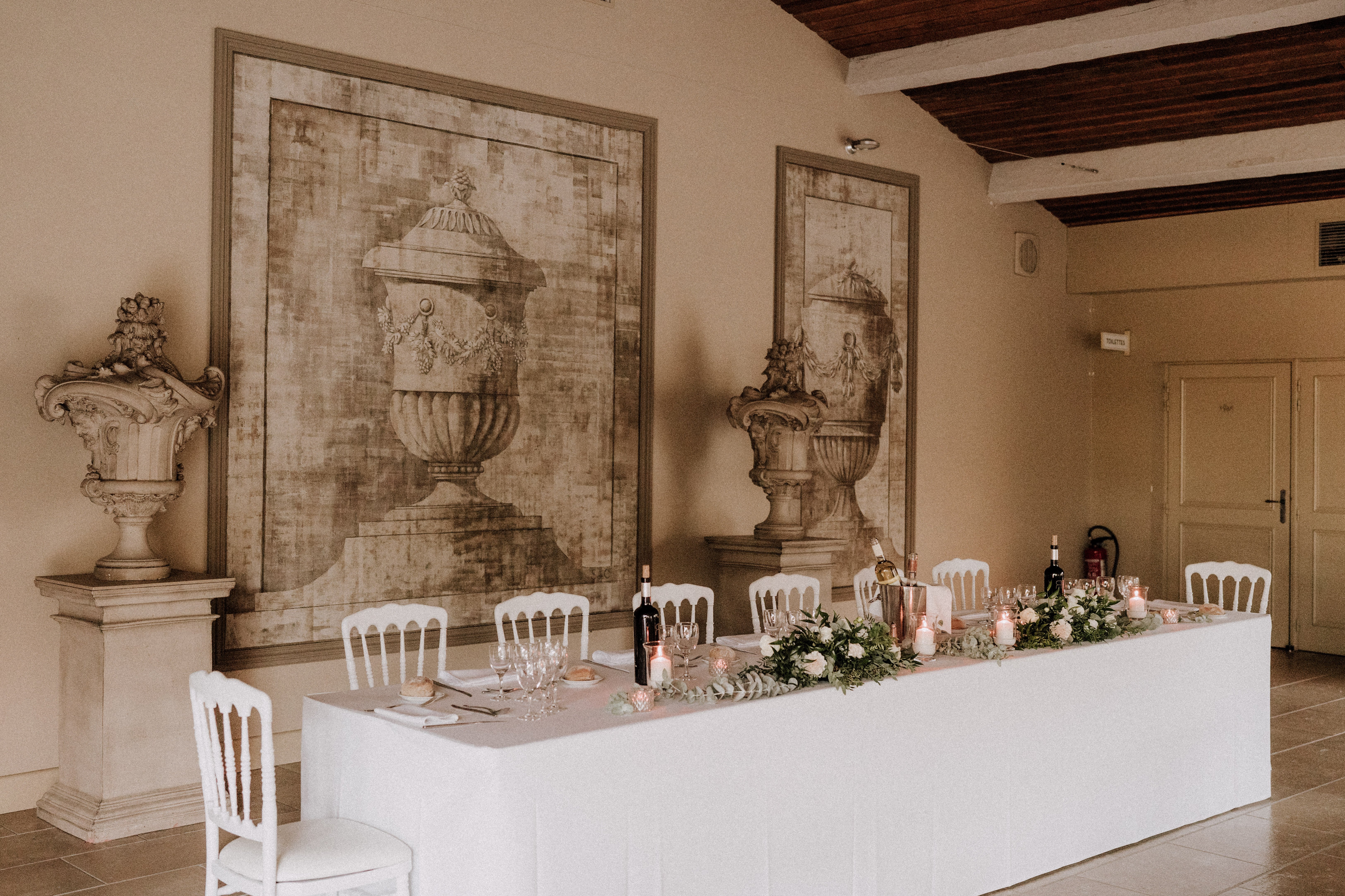 A wedding reception head table is set up indoors in a classic French venue with warm beige walls, exposed dark wood ceiling beams, and large-scale sepia-toned trompe-l'œil murals depicting ornamental urns. The long rectangular table is covered in a white linen and seats approximately eight guests using white Napoleon-style chairs. The centerpiece features a garland of green eucalyptus and foliage interspersed with white and blush roses, pillar candles in varying heights, and small votive candles providing warm ambient lighting. Wine bottles, a champagne bucket, crystal glassware, and folded napkins are placed at each setting, and a decorative stone urn sculpture sits on a pedestal to the left. The overall decor palette is white, blush, and green against warm neutral tones, with a classic French chateau styling. Wide shot taken from a slight angle. Potential venue feature image.