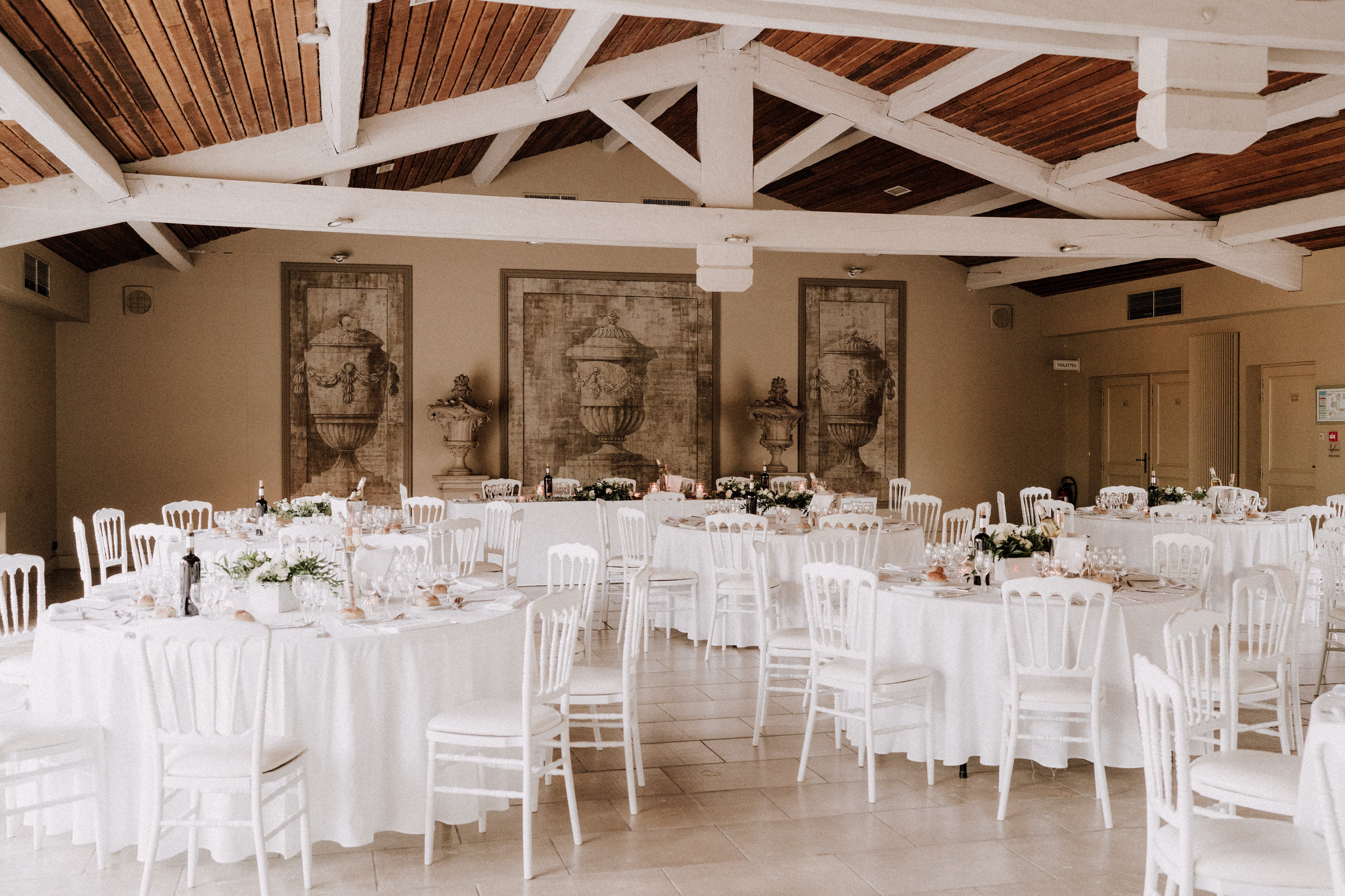 A wide shot of an indoor wedding reception space set up for a seated dinner, with no guests present. The room features a vaulted ceiling with white-painted timber trusses and warm dark wood planking, along with a light stone tile floor. Three large trompe-l'oeil mural panels depicting classical urns are painted on the back wall in muted sepia tones. Multiple round tables are dressed in white linen tablecloths and surrounded by white Napoleon-style chairs; tables are set with glassware, plates, and low centerpieces of white and green florals with small candles and wine bottles. The overall decor palette is white and ivory with soft green accents, giving the space a classic, clean aesthetic. Potential venue feature image.