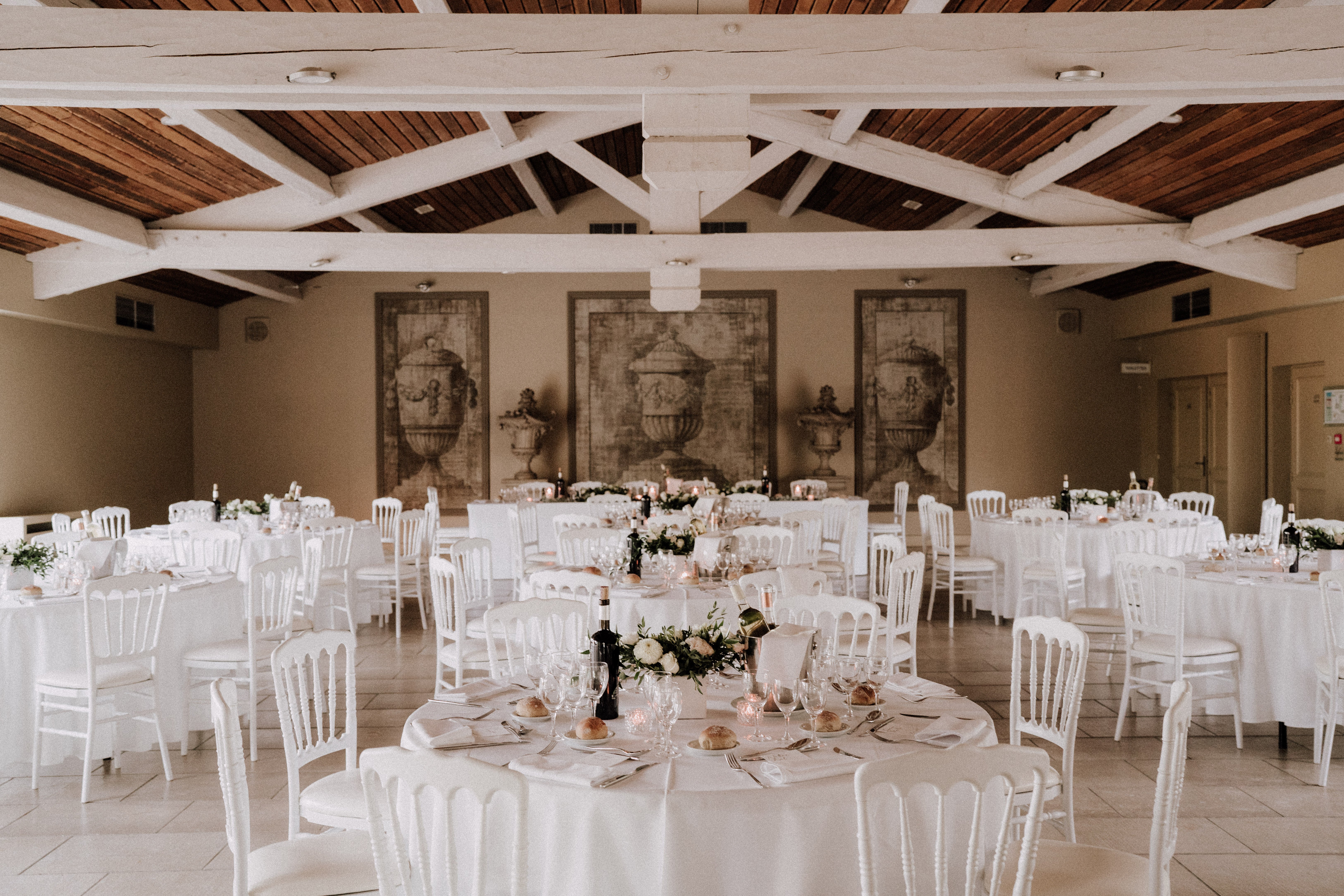 A wide shot of an indoor wedding reception hall set up for dinner, with no guests present. The room features a vaulted ceiling with white-painted wooden beams and dark wood planking, giving it a classic French country character. Multiple round tables are dressed in white floor-length linens and surrounded by white Napoleon-style chairs, with the foreground table displaying a low centerpiece of white roses, blush blooms, and green foliage alongside small votive candles, wine glasses, bread rolls, and dark wine bottles. Three large sepia-toned decorative panels depicting ornate urns are mounted on the back wall, anchoring the room's neutral, classic decor palette of white, ivory, and warm beige. Potential venue feature image.