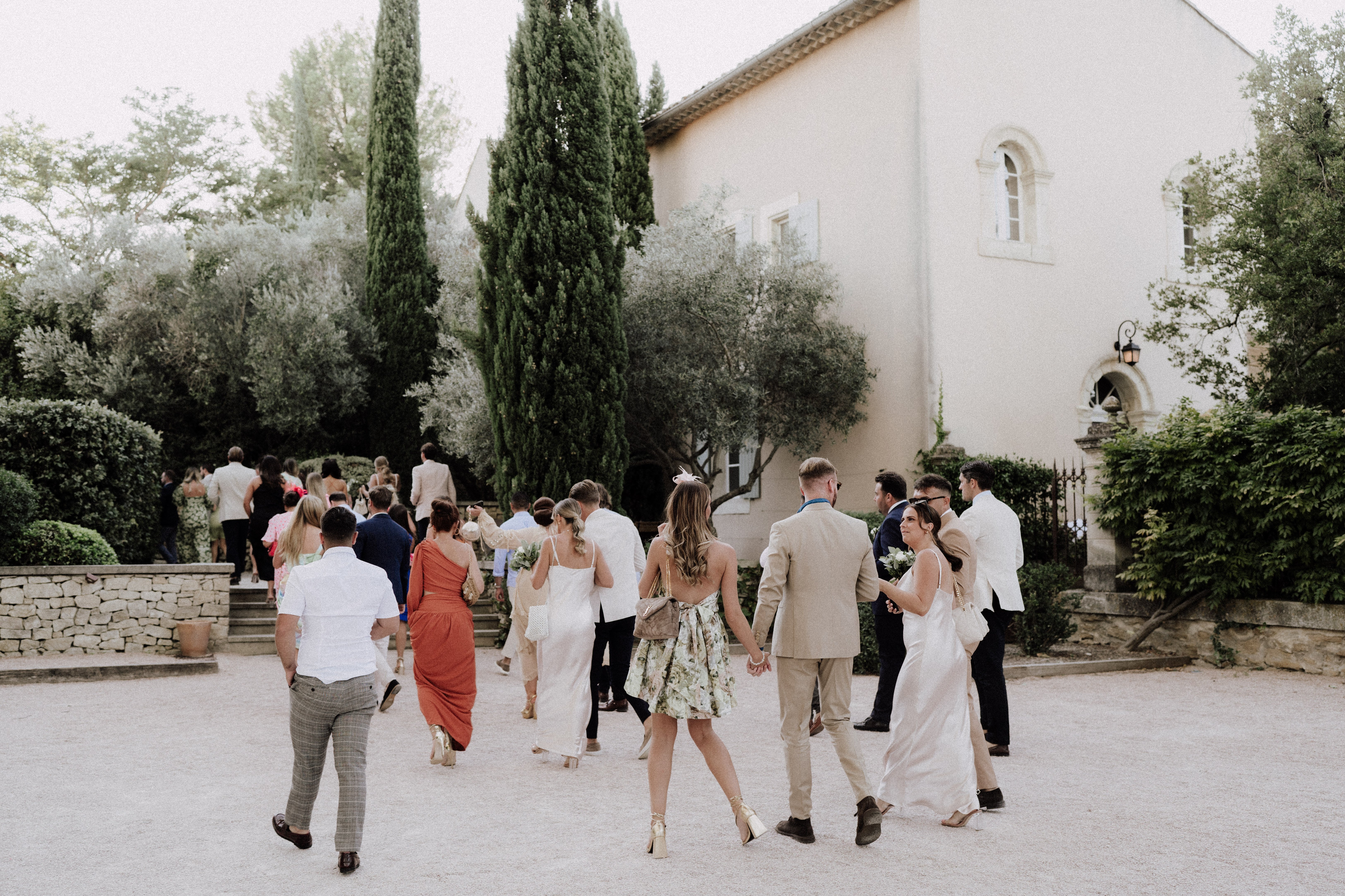 A group of approximately 20 wedding guests is walking across a gravel courtyard toward a classic Provençal-style manor house with cream-rendered walls, arched windows, and a terracotta tile roof. The scene appears to be during a cocktail hour or guest arrival, with people moving toward stone steps leading to the entrance. Guests are dressed in a mix of summer wedding attire including an rust-orange one-shoulder maxi dress, a floral mini dress paired with gold heeled sandals, a white slip dress, a beige linen suit, navy suits, and a white blazer. The composition is a wide candid shot taken from behind the group, giving a sense of movement and arrival at the venue. Potential venue feature image.