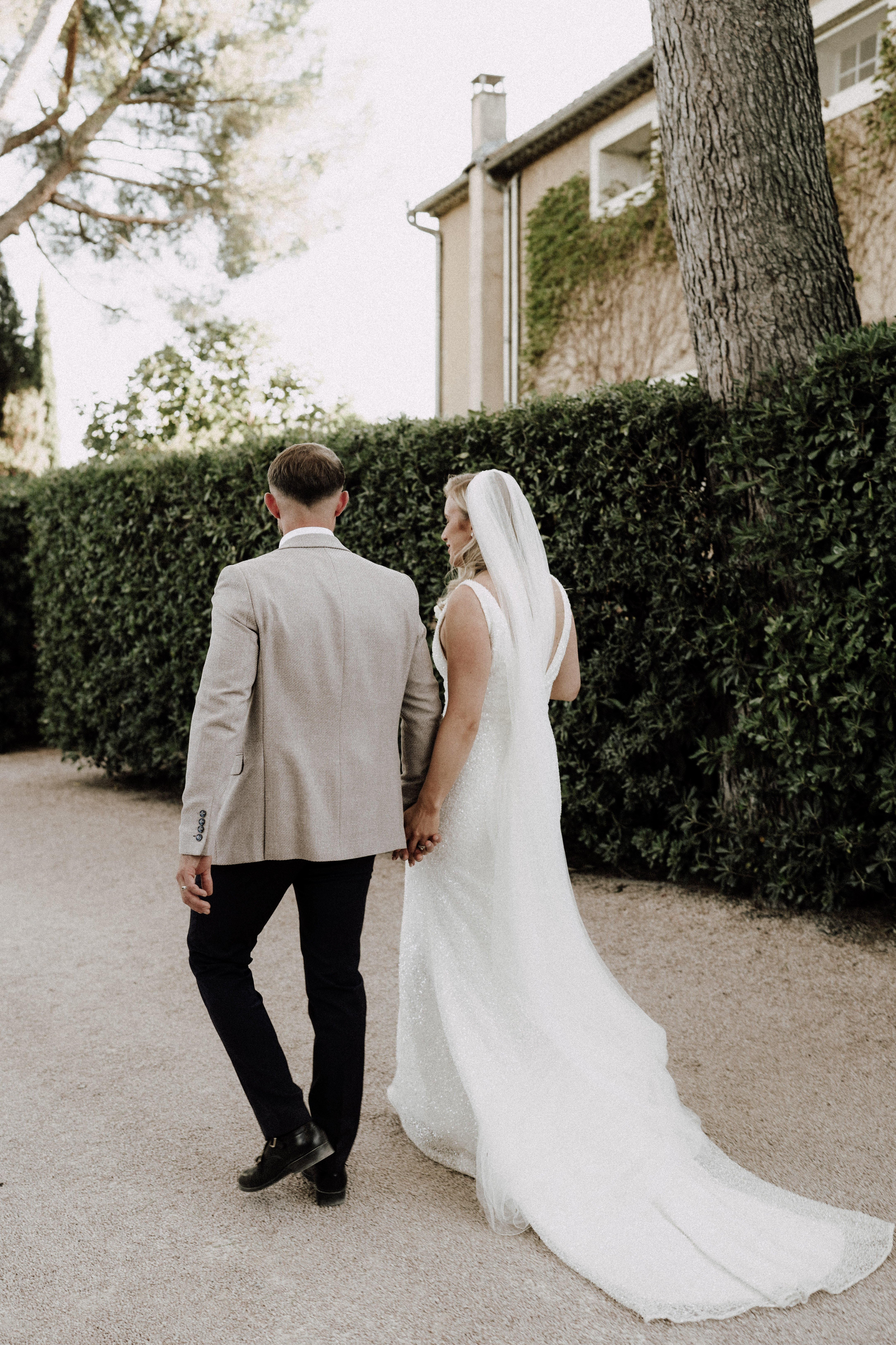 A couple portrait taken from behind as the bride and groom walk hand-in-hand along a gravel path beside a neatly trimmed tall hedge, with a stone manor building partially visible in the background. The groom wears a light taupe blazer with dark trousers and black leather shoes, while the bride wears a sleeveless, low-back white fitted gown with a subtle shimmer texture and a long cathedral-length train, paired with a shoulder-length veil. The styling is clean and modern with a neutral palette. The shot is a full-length wide portrait composition captured from the back.