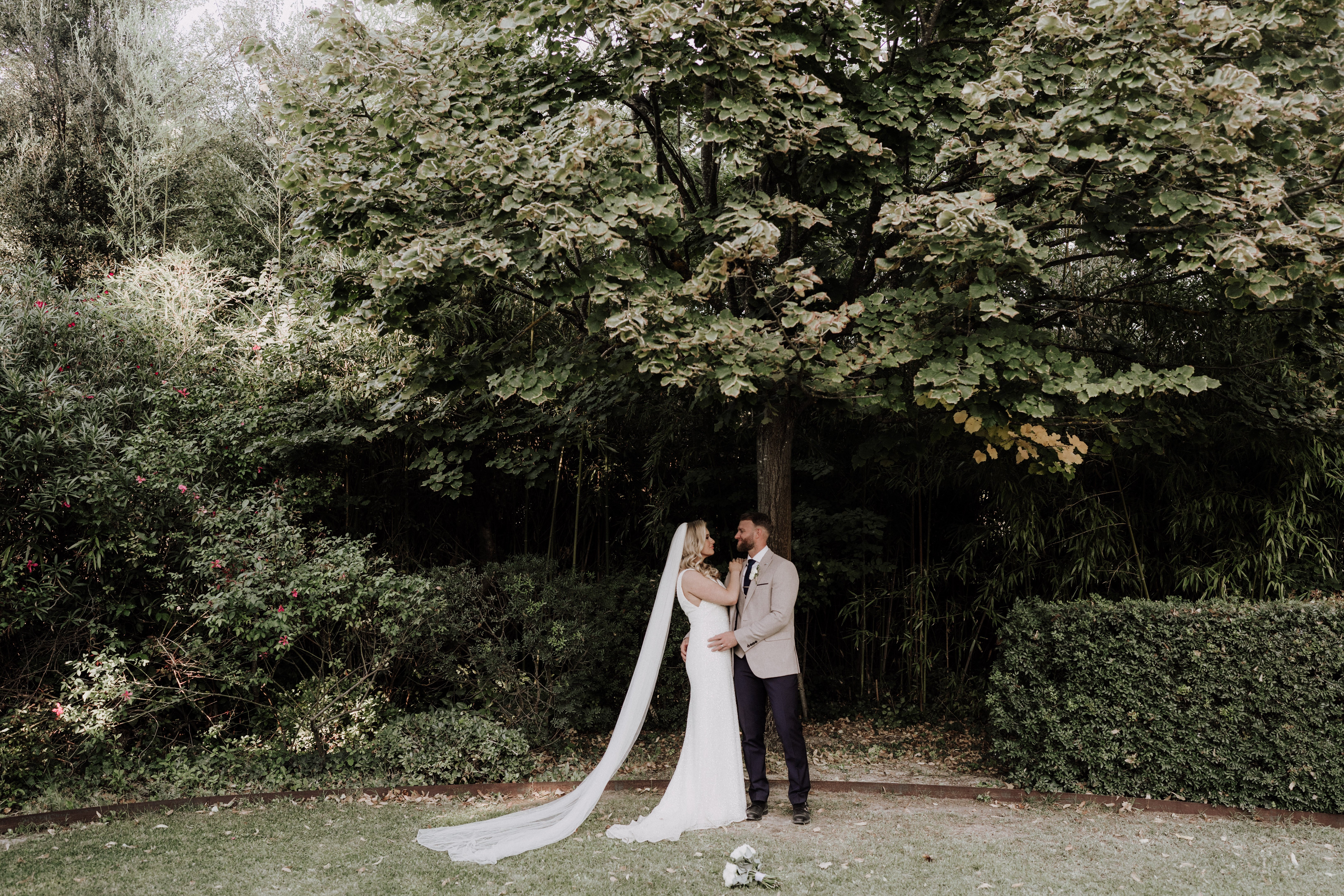 A couple portrait taken outdoors in a garden setting, with the bride and groom standing close together beneath a large tree, facing each other. The bride wears a fitted white gown with a long cathedral-length veil that trails across the lawn, and her bouquet of white flowers rests on the ground nearby. The groom is dressed in a beige blazer, navy trousers, and a dark tie. The composition is a wide shot that places the couple small within the frame, emphasizing the lush garden surroundings including manicured hedges, bamboo, and shrubs with small red flowers.