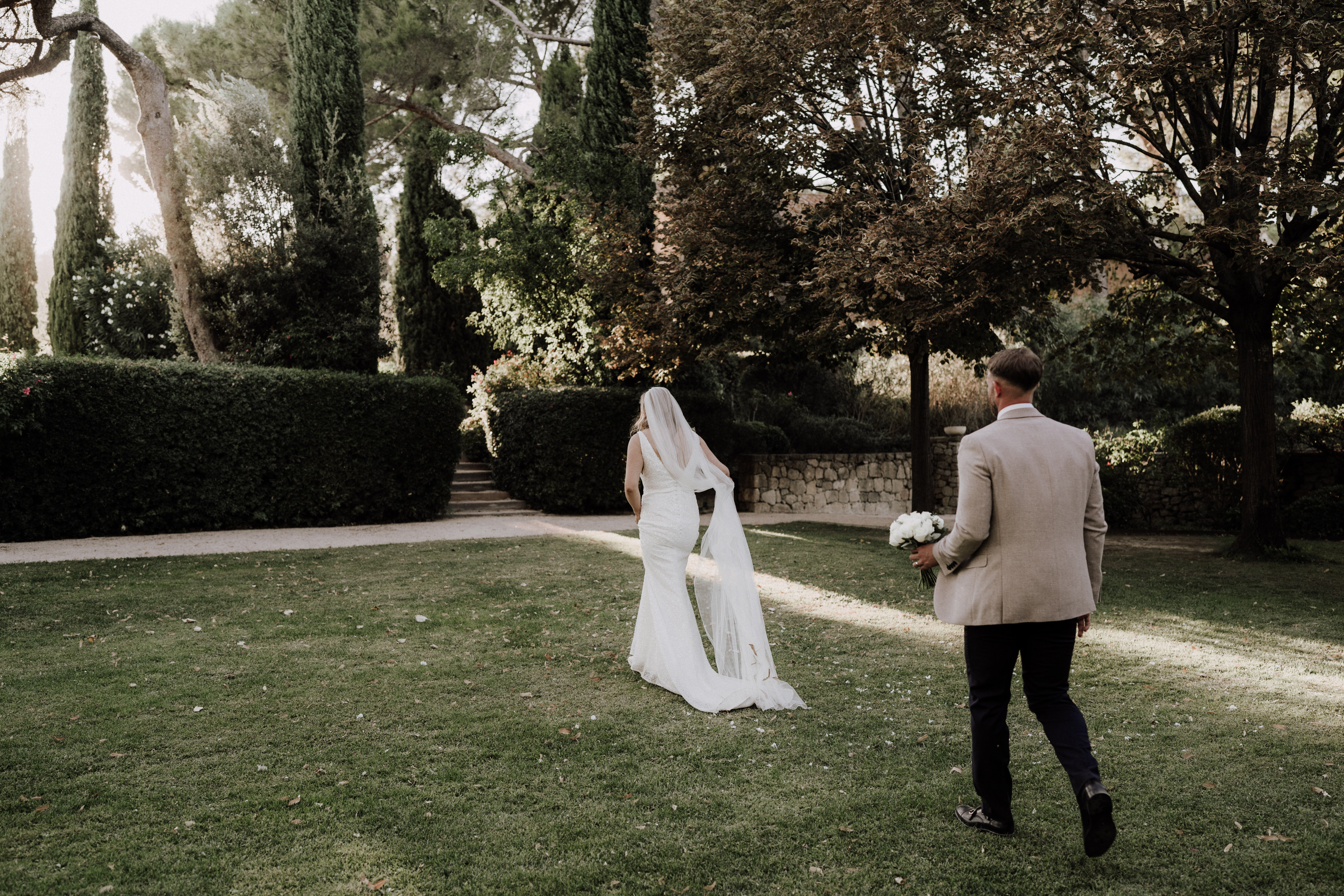 The bride and groom are walking together across a manicured lawn in an outdoor garden setting, photographed from behind in a wide shot. The bride wears a fitted white gown with a long cathedral-length veil, while the groom is dressed in a sand-colored blazer with dark trousers; he carries a bouquet of white flowers, likely peonies. The garden features trimmed hedgerows, cypress trees, and a low stone wall with steps leading to a pathway, suggesting a formal French estate or chateau grounds. The overall styling is classic and minimal, with warm late-afternoon light casting long shadows across the lawn.