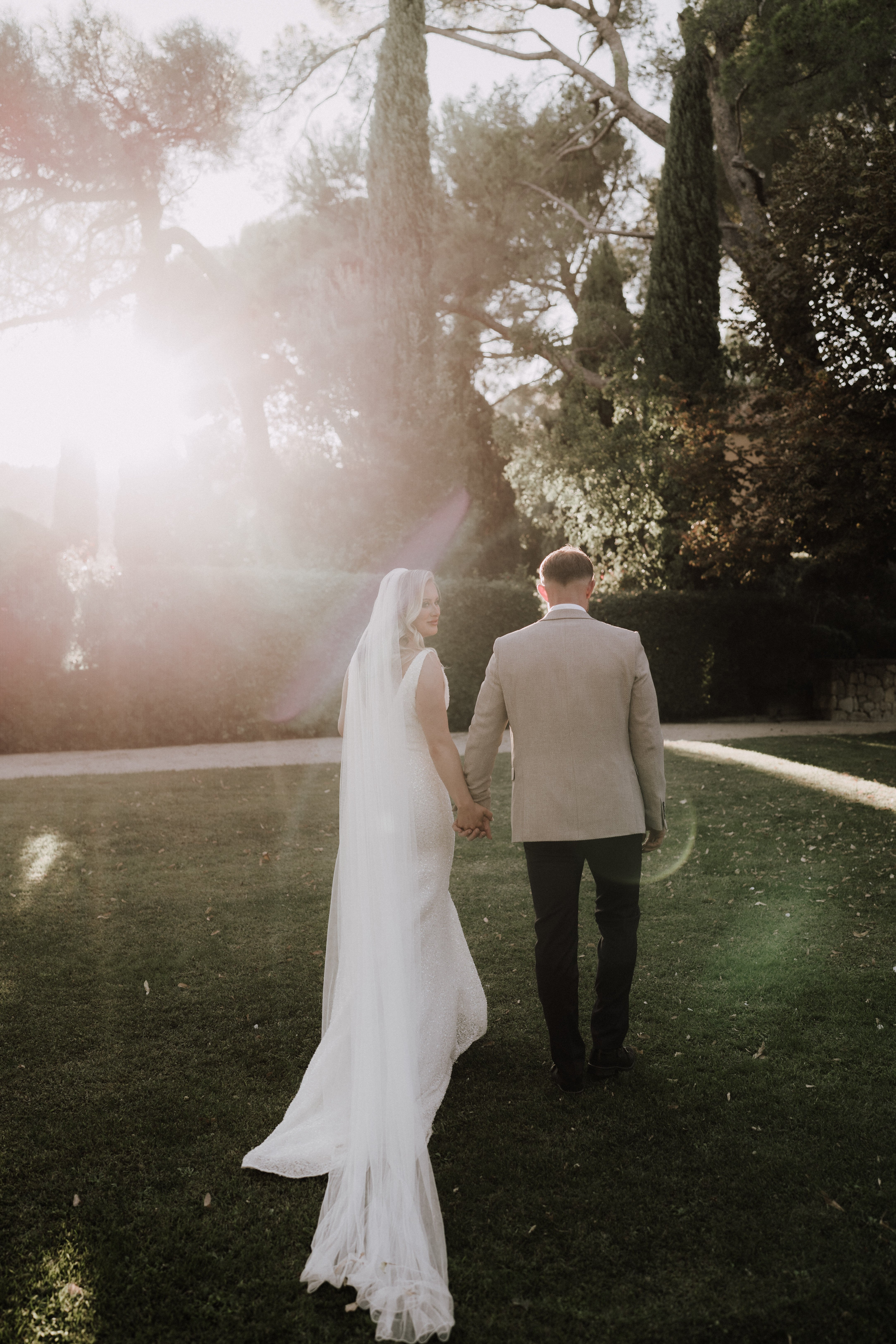 A couple portrait taken outdoors in a formal garden setting, with the bride and groom walking away from the camera while holding hands. The bride glances back over her shoulder and wears a fitted white lace gown with a long train and a cathedral-length plain veil, while the groom wears a tan/champagne blazer with dark trousers. The shot is taken during golden hour, with strong lens flare from the low sun creating a warm glow across the upper left of the frame. Tall cypress trees and manicured hedges frame the background. The composition is a full-length rear portrait with the couple centered on a well-kept lawn.