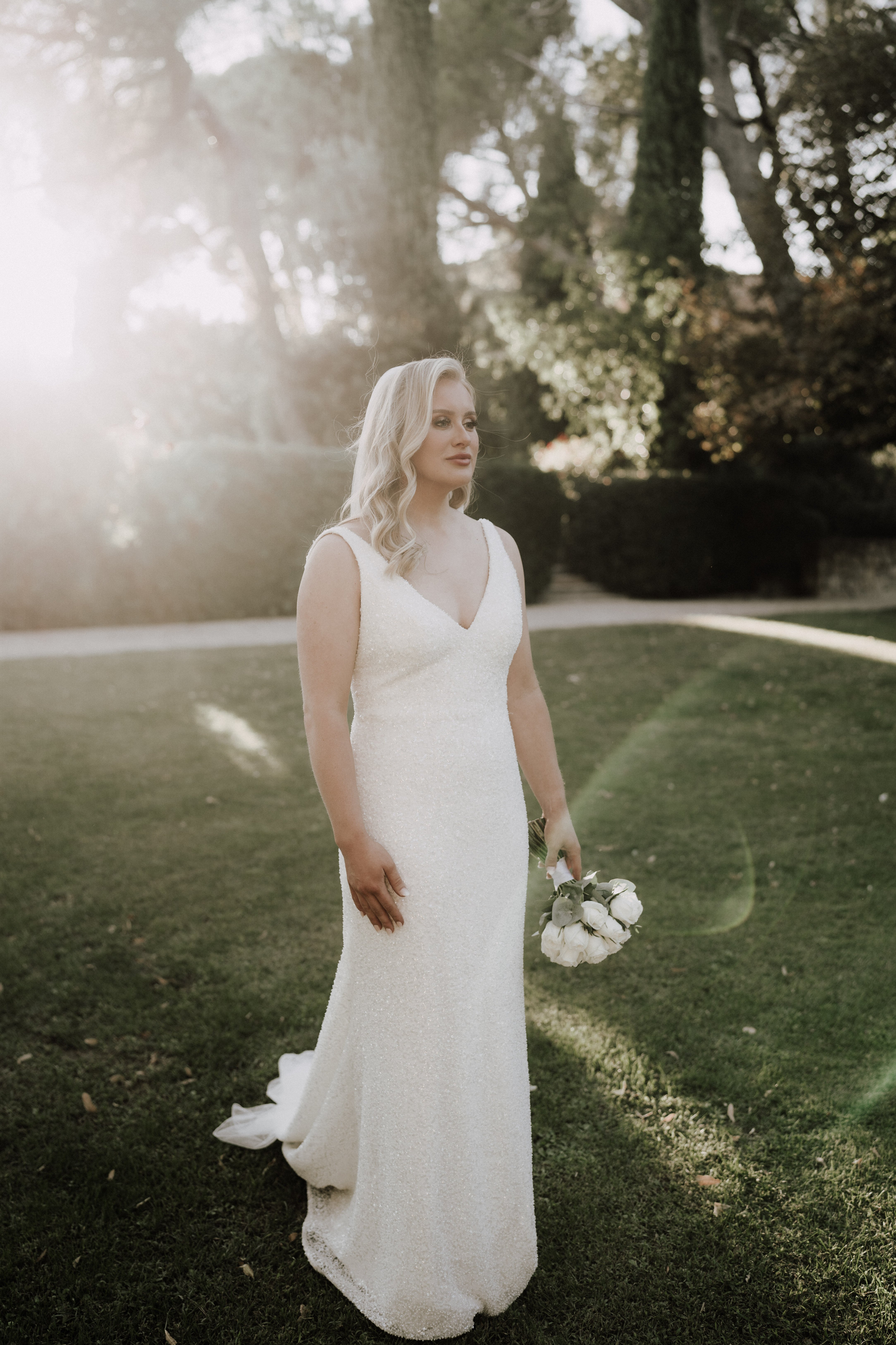 A bridal portrait taken outdoors on a manicured lawn, with the bride standing alone and gazing to one side. She wears a fitted, sleeveless ivory sequined gown with a V-neckline and a short train, and holds a small, loosely gathered bouquet of white peonies with minimal greenery at her side. Her blonde hair is styled in loose waves, and warm golden backlight creates lens flare across the left side of the frame. The background features trimmed hedgerows and tall mature trees, suggesting formal garden grounds. The shot is a full-length portrait with a slightly warm, sun-drenched tone.
