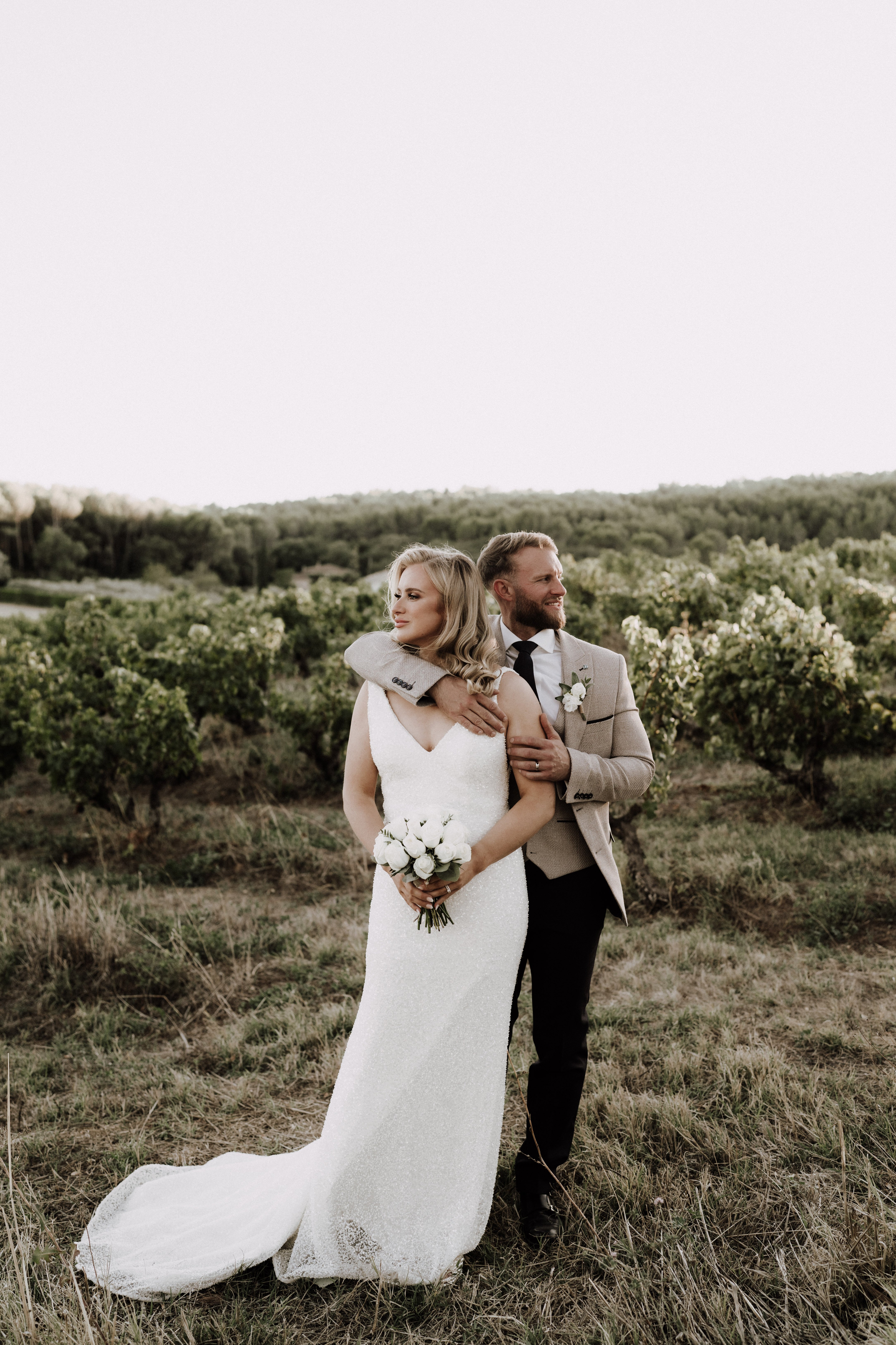 An outdoor couple portrait taken in a vineyard setting, with rows of grapevines stretching across the background. The bride wears a fitted ivory lace gown with a V-neckline and a trailing train, holding a compact bouquet of white garden roses with minimal greenery; the groom's tan blazer is draped over her shoulders. The groom is dressed in dark trousers, a white shirt, black tie, and a white floral boutonniere, and stands behind the bride with his hands on her arms as both look off to the side. The composition is a full-length portrait shot with a clean, muted color palette emphasizing the neutral tones of the couple's attire against the vineyard landscape.