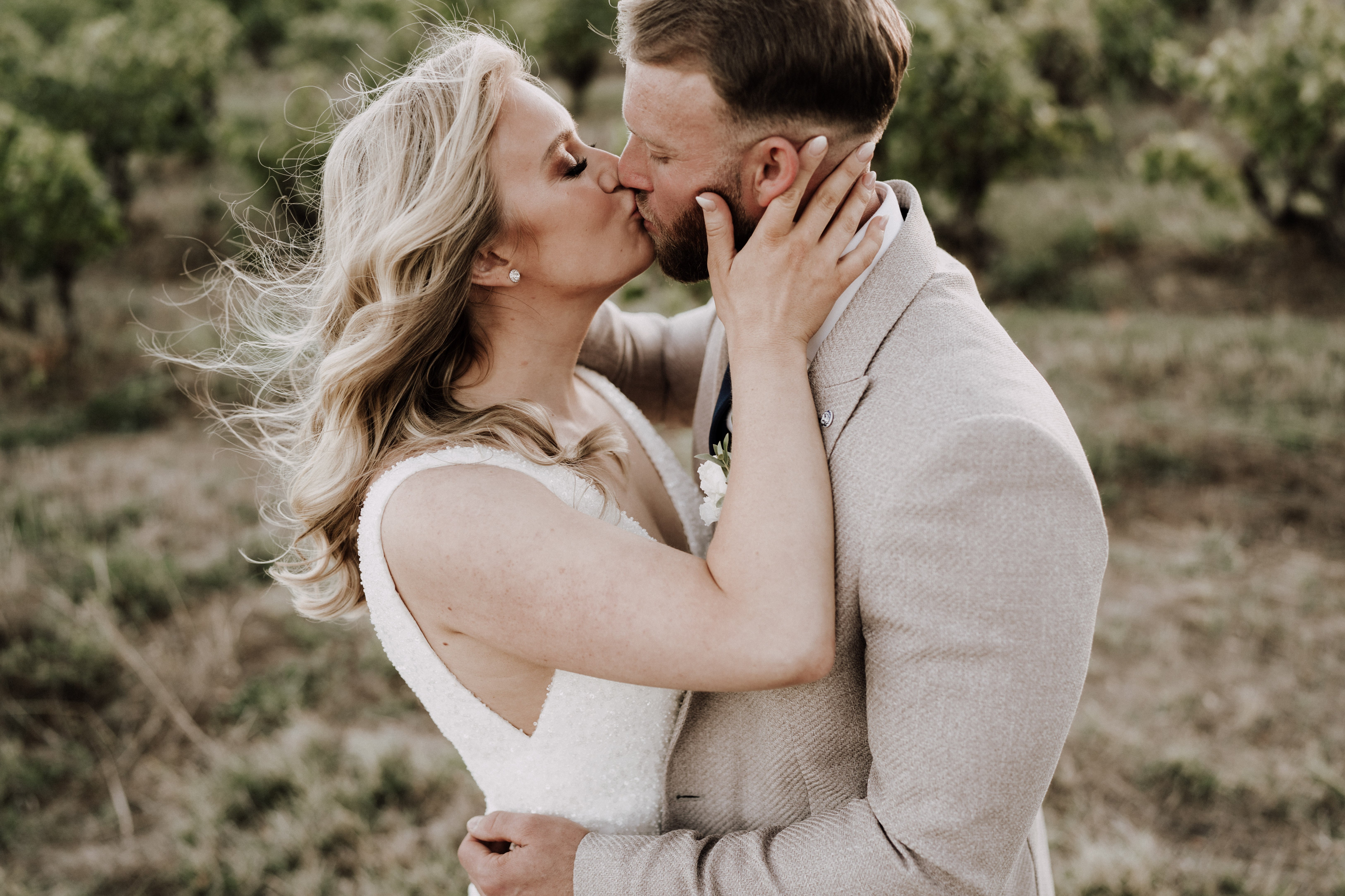 A couple portrait taken outdoors in what appears to be an orchard or vineyard setting, with the bride and groom kissing. The bride wears a sleeveless white beaded gown with a sweetheart neckline and has long blonde hair, windswept to one side; she wears small diamond stud earrings. The groom is dressed in a light taupe suit with a white floral boutonniere and a navy pocket square or tie visible at the collar. The composition is a medium close-up shot with a shallow depth of field, keeping the couple sharp against a softly blurred green orchard background. The overall styling is clean and modern with a neutral color palette.