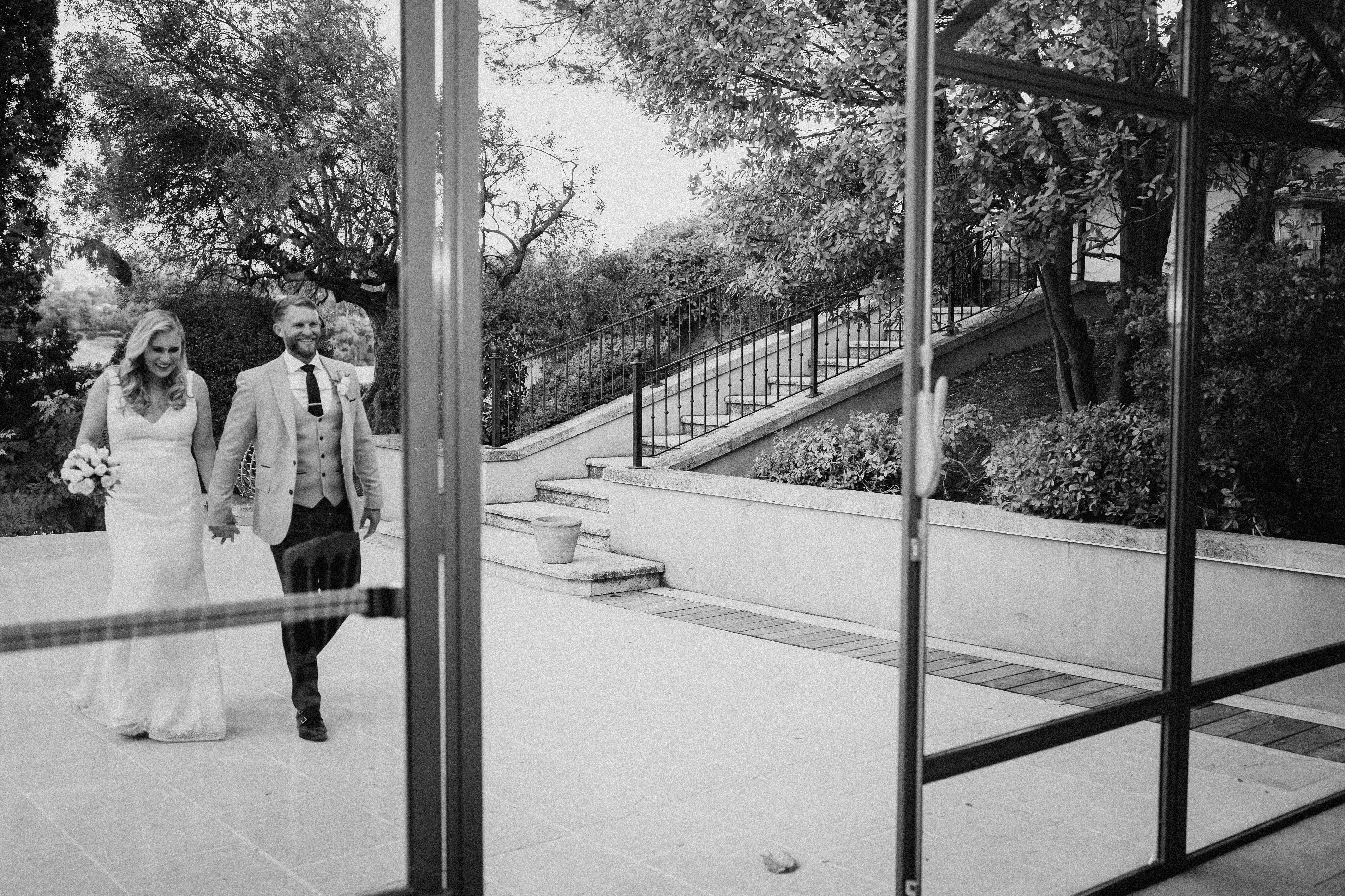This black-and-white couple portrait shows a bride and groom walking hand-in-hand across a stone terrace, photographed through large glass doors with dark metal frames. The bride wears a fitted, sleeveless gown and carries a round bouquet, while the groom wears a light-toned suit with a waistcoat, dark tie, and a buttonhole flower. Both are smiling and relaxed. The outdoor setting features a formal terrace with stone steps, wrought-iron railings along a sloped staircase, and terracotta planters. The image is rendered in high-contrast black and white with soft mid-tones, and the creative composition frames the couple through the reflective glass doors, placing them slightly left of center within a wide environmental shot.