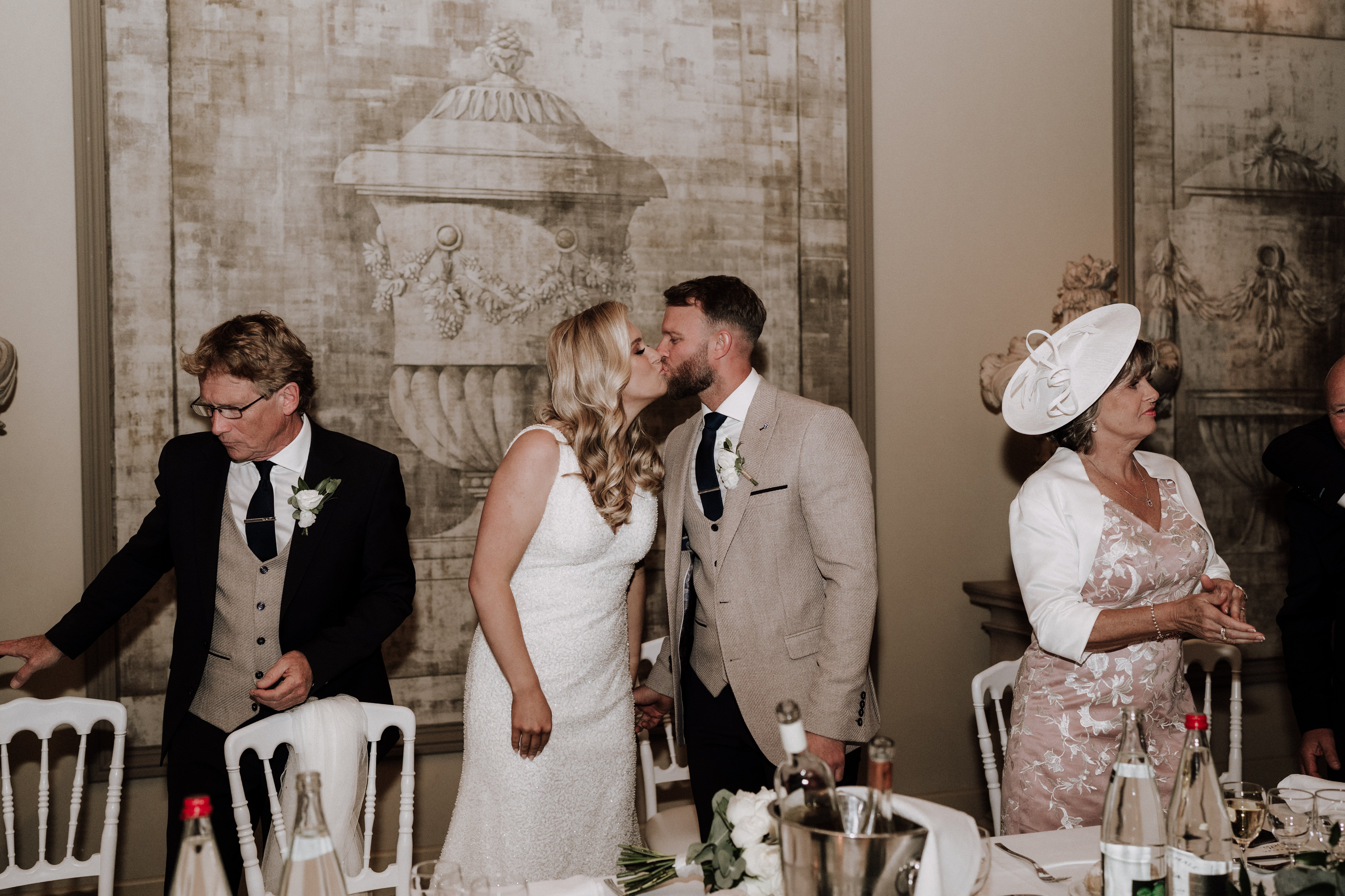 The bride and groom share a kiss during what appears to be the wedding reception, standing at the top table in a formal indoor room. The bride wears a sleeveless fitted white textured gown with long blonde wavy hair, and the groom is dressed in a tan/beige tweed three-piece suit with a navy tie and a white buttonhole flower. To the left stands an older man in a dark suit with a beige waistcoat and navy tie, also wearing a white buttonhole, and to the right a woman in a dusty pink floral dress with a white bolero jacket and a large white fascinator hat. The room features large-scale classical trompe-l'oeil mural panels depicting urns and architectural motifs in neutral grey-beige tones, and the table in the foreground is set with white linens, white Chiavari chairs, champagne bottles in an ice bucket, glassware, and a low white floral centerpiece. Wide mid-shot composition capturing four people at the top table.