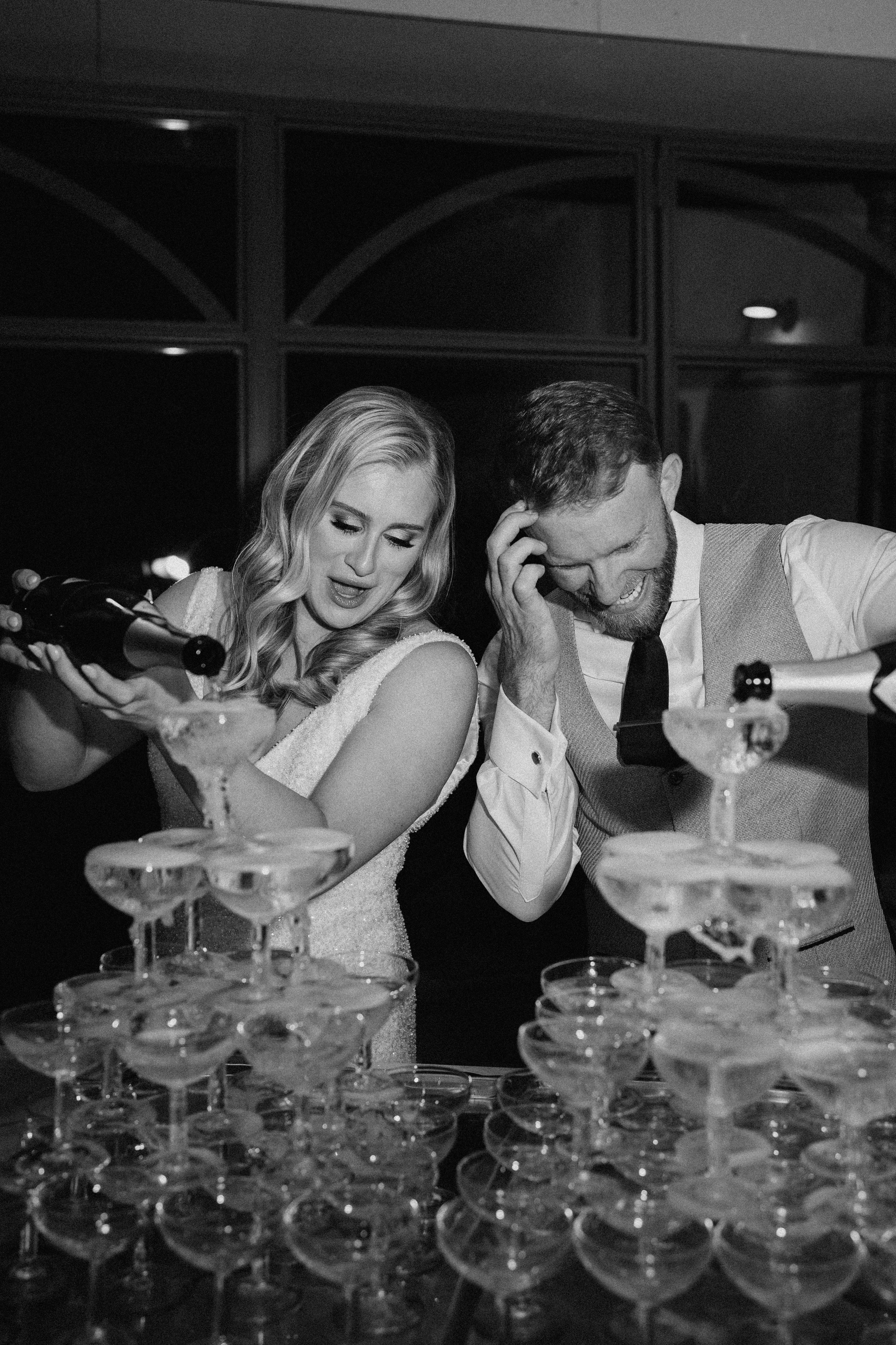 This black-and-white image captures the bride and groom pouring champagne into two large coupe glass tower pyramids during an indoor evening reception. The bride, wearing a short-sleeved sequined or beaded white dress with loose waves in her hair, leans forward concentrating as she pours from a bottle, while the groom — dressed in a vest, tie, and white dress shirt with rolled sleeves — laughs and covers his face with his hand in an amused reaction. The high contrast between the dark background and the bright whites of their attire and glassware gives the image a dramatic, editorial feel. The setting appears to be an indoor venue with large arched windows visible in the background, and the composition is a close-up portrait shot of the couple from the waist up.
