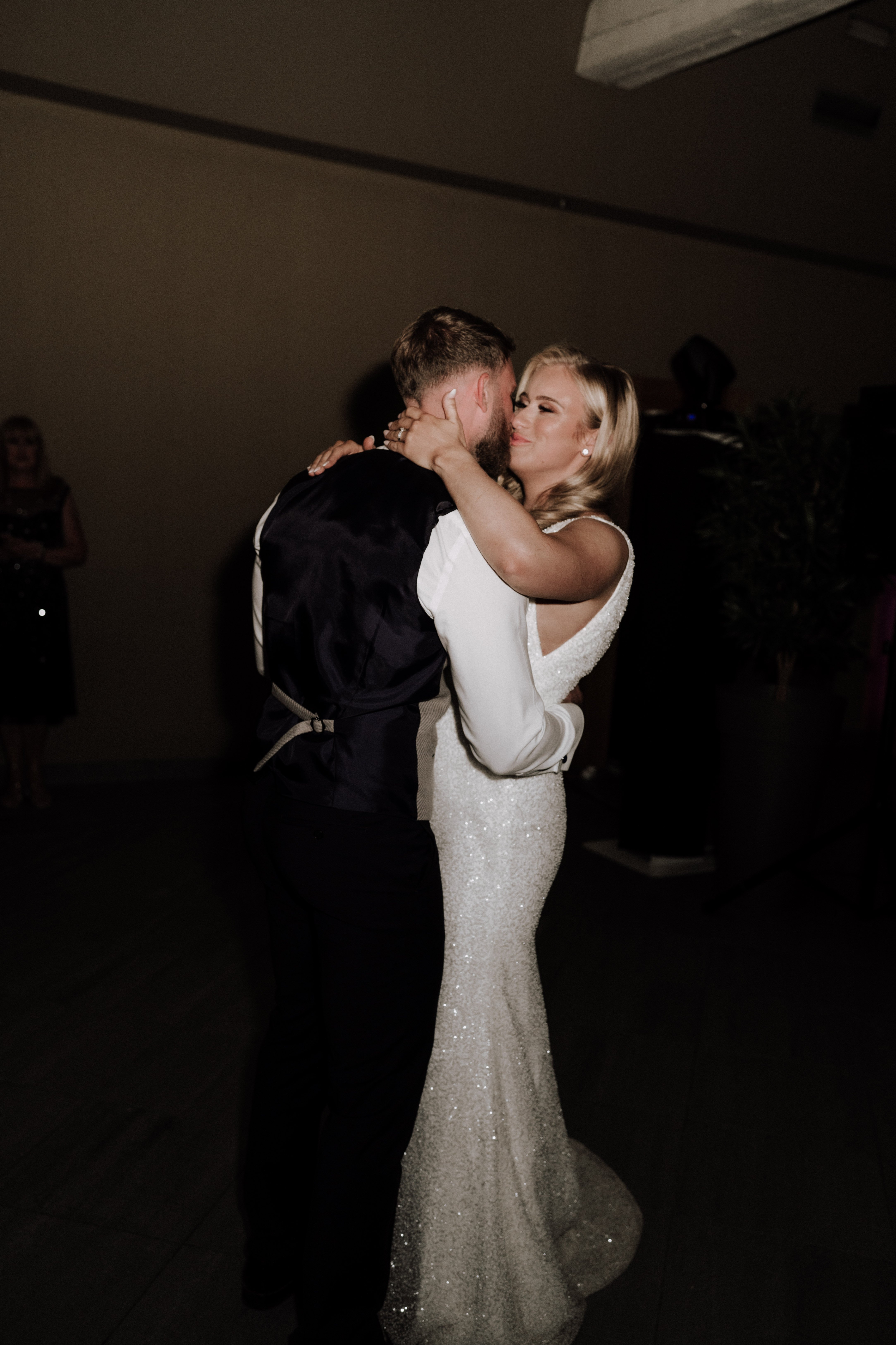 The bride and groom share their first dance in a dimly lit indoor reception space, with a small number of guests visible in the dark background. The bride wears a form-fitting ivory sequined gown with long sleeves and an open back, her blonde hair styled down, and she has her arms wrapped around the groom's neck. The groom is dressed in a dark navy suit with a white waistcoat. The overall styling is modern and classic, with low ambient lighting creating a dark, moody atmosphere. Medium portrait shot taken at floor level, centered on the couple.