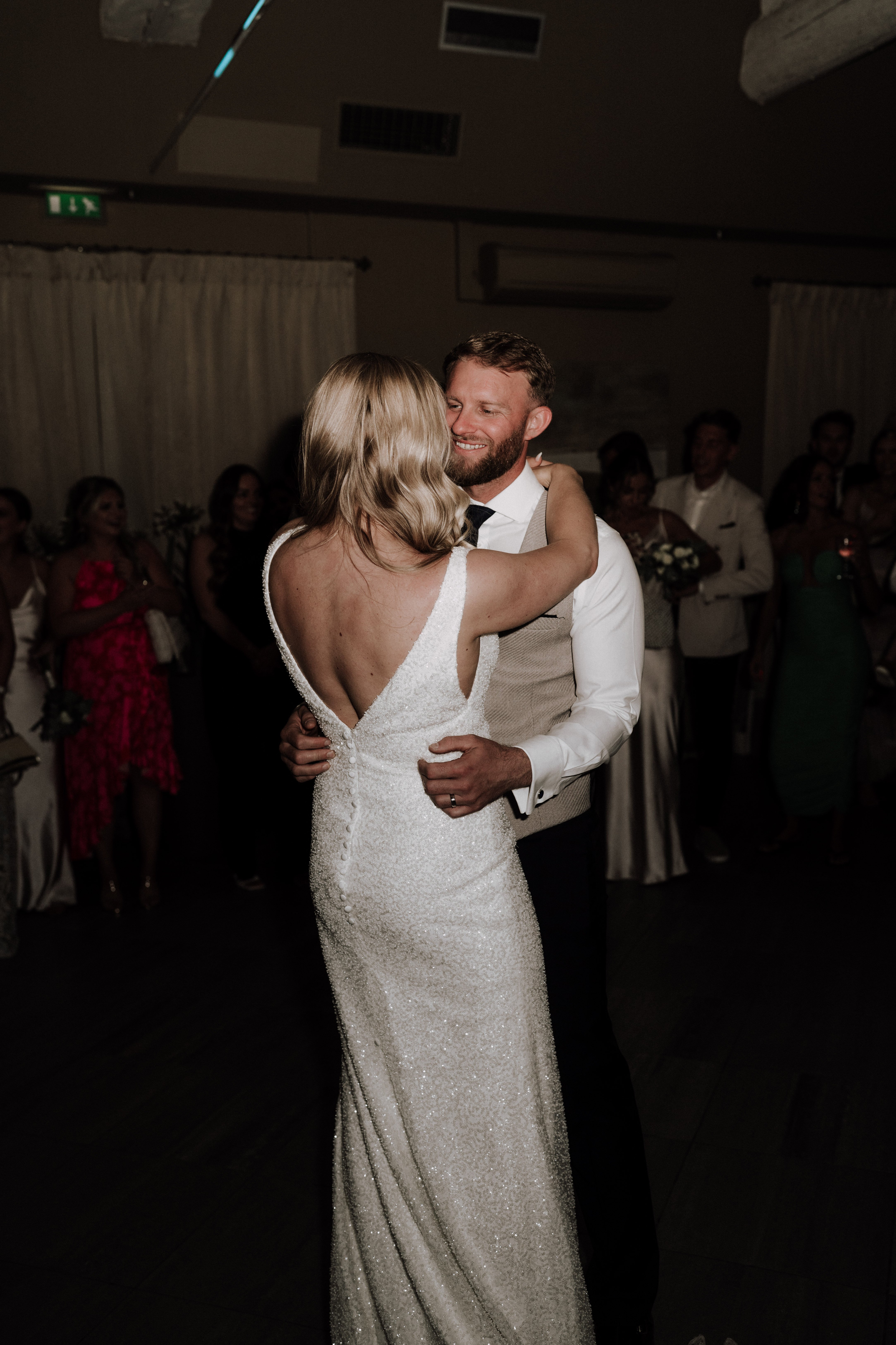 The bride and groom share their first dance in a dimly lit indoor reception room, surrounded by a circle of approximately 15-20 guests watching from the edges of the dance floor. The bride wears a form-fitting, fully beaded ivory gown with a deep V-back, button detail along the spine, and thin straps, paired with loose blonde waves; the groom wears a white dress shirt, dark tie, and a beige textured waistcoat with dark trousers. The groom is smiling broadly as he holds the bride close, his hand placed on her lower back. The shot is taken from behind and slightly to the side of the bride, framing the couple in a medium portrait composition with guests visible in the soft background, some in bright pink and white outfits.