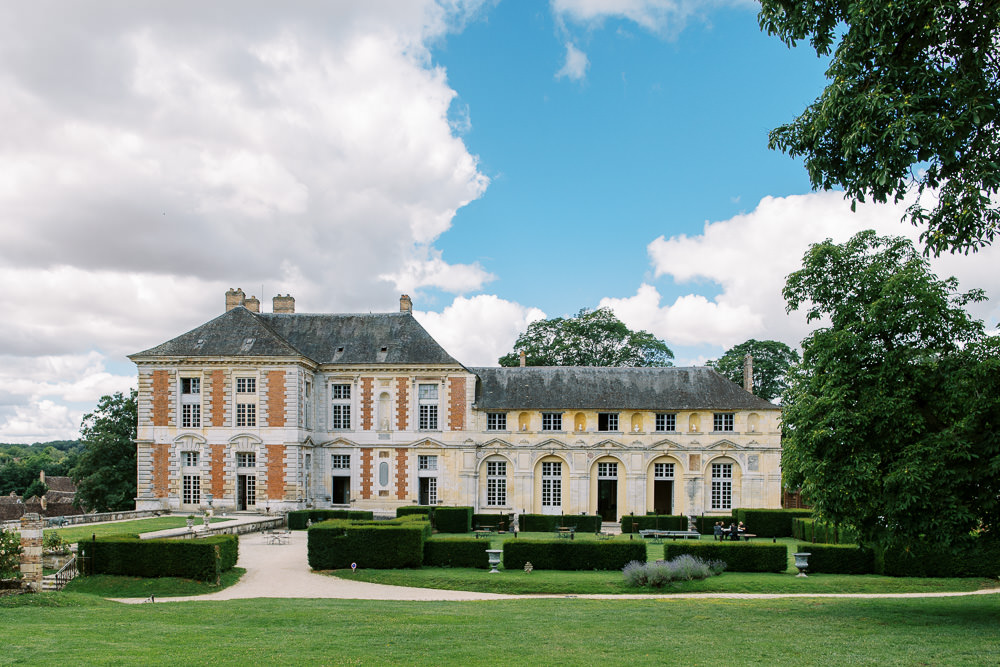 Wide exterior shot of a large French classical chateau featuring cream stone and red brick facade with mansard slate roofs, tall arched windows, and ornamental stonework detailing across multiple wings. The formal grounds in the foreground include neatly clipped boxwood hedges arranged in a geometric parterre pattern, gravel pathways, and decorative stone urns. A small number of people are visible seated near the hedgerows, likely guests. No wedding party or ceremony activity is visible; this is primarily a venue establishing shot taken in daylight. Potential venue feature image.