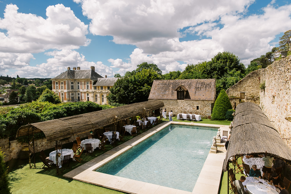 An elevated wide shot of an outdoor cocktail hour or reception setup at a French chateau estate. A rectangular pool sits at the center of the courtyard, flanked on the left by a long pergola structure with a thatched or reed roof sheltering multiple round tables dressed in white linens, surrounded by wooden folding chairs. On the right side, a smaller thatched-canopy structure covers additional seating where guests are visible. A handful of guests and staff are scattered around the pool area. In the background, a grand French classical chateau with pale yellow rendered facades, tall mansard-style chimneys, and large windows is visible, along with a stone barn building and clipped conical topiary. The overall decor palette is neutral — white table linens, natural wood and thatched structures — set against the historic stone walls of the estate grounds. Potential venue feature image.