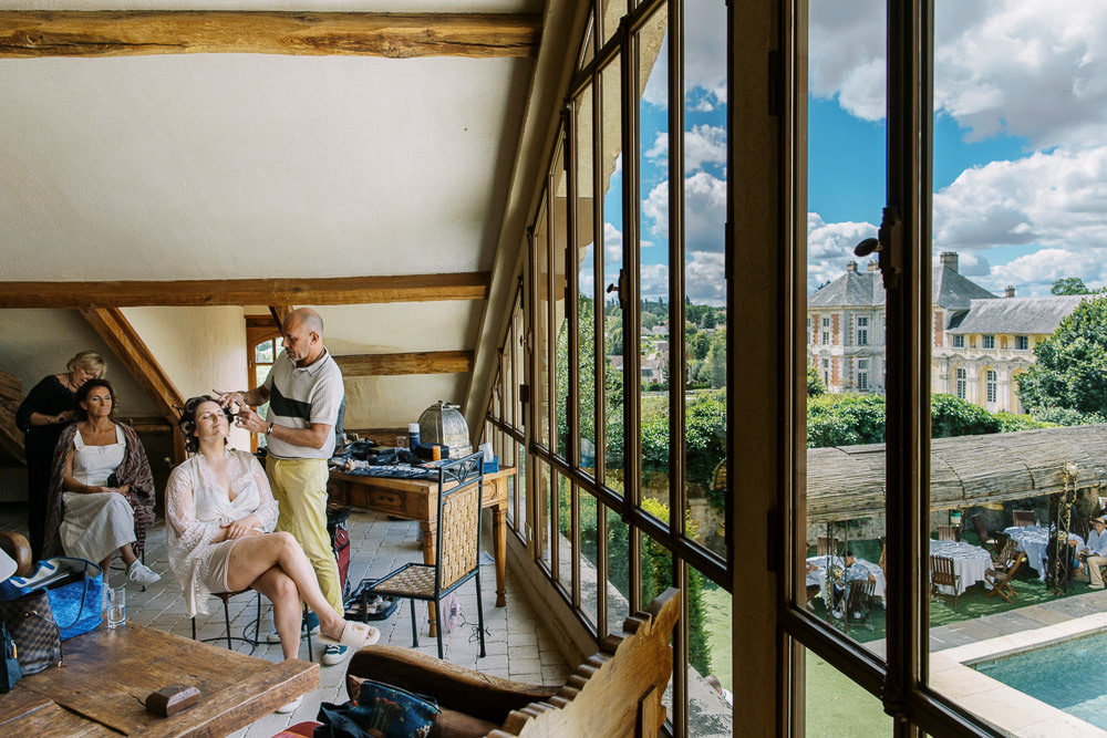 A getting-ready scene taking place in a rustic attic-style room with exposed wooden beams and large floor-to-ceiling steel-framed windows. A bride seated in a chair is having her hair styled by a male hairstylist wearing a white and grey polo shirt and yellow trousers, while another woman in white sits nearby having her makeup done by a second attendant. The room has a casual, lived-in feel with makeup bags, a Louis Vuitton tote, and styling tools spread across wooden furniture. Through the large windows, a classical French chateau is clearly visible in the background, along with a courtyard below where white-clothed reception tables and guests are already gathering around a pool area. The composition is a wide interior shot that uses the window as a framing device to simultaneously capture the getting-ready moment and the venue exterior. Potential venue feature image.