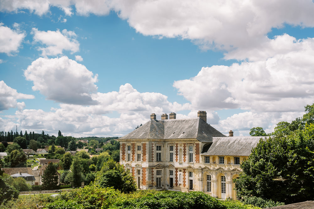 Wide exterior shot of a French château taken from an elevated vantage point, showing the full façade of a grand 17th or 18th-century classical building with cream and red brick stonework, tall arched windows on the ground floor, multiple dormer windows set into the steep slate mansard roof, and several chimney stacks. The château is partially surrounded by mature trees and shrubs, with a small French village and rolling countryside visible in the background. No people are present in the frame. Potential venue feature image.