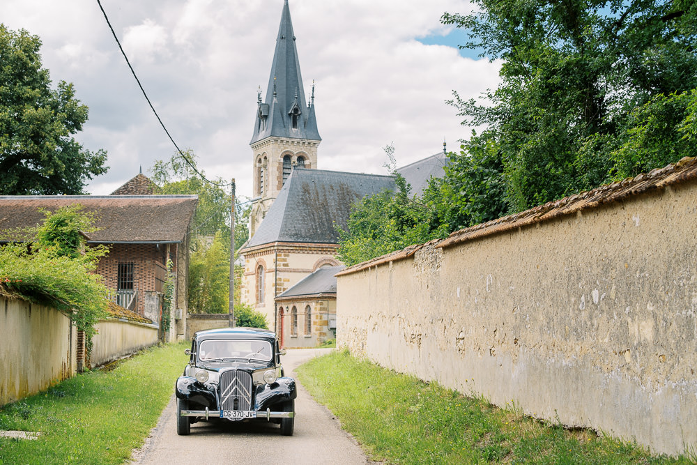 A classic black Citroën Traction Avant wedding car drives along a narrow village lane toward the camera, with a French rural church featuring a pointed slate steeple visible in the background. The setting is an outdoor French village, with a rendered stone boundary wall running along the right side of the lane and a brick outbuilding to the left. The vintage black car serves as the wedding transport, adding a retro styling element to the day. Wide shot composition capturing both the vehicle and the village architectural context.