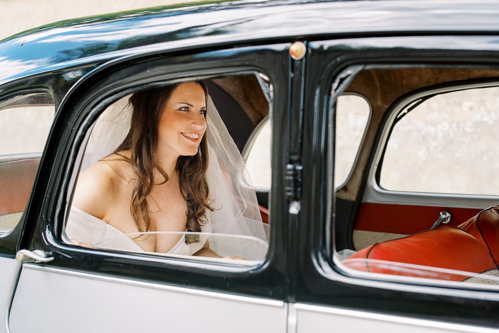 A bride is seated inside a vintage two-tone black and white car with a red leather interior, photographed through the open rear window. She is wearing an off-the-shoulder white dress and a long flowing veil, with her dark brown hair worn down in loose waves. She is smiling and looking to one side, suggesting she may be arriving at the ceremony. The shot is a medium portrait framed by the car's window and door trim, with the retro vehicle serving as a strong compositional element.