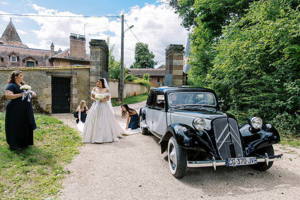 The bride stands outdoors on a gravel driveway in front of what appears to be a French chateau or manor entrance with stone gate pillars and brick outbuildings, holding a bouquet of white flowers. She is wearing an off-the-shoulder white ball gown with a long train, which two bridesmaids dressed in floor-length navy blue gowns are crouching down to arrange. A third bridesmaid in a matching navy dress stands to the left also holding a white bouquet. A classic black Citroën Traction Avant vintage car is parked prominently to the right of the frame. The image is a wide shot taken in natural daylight, capturing a pre-ceremony moment as the bridal party prepares for departure.