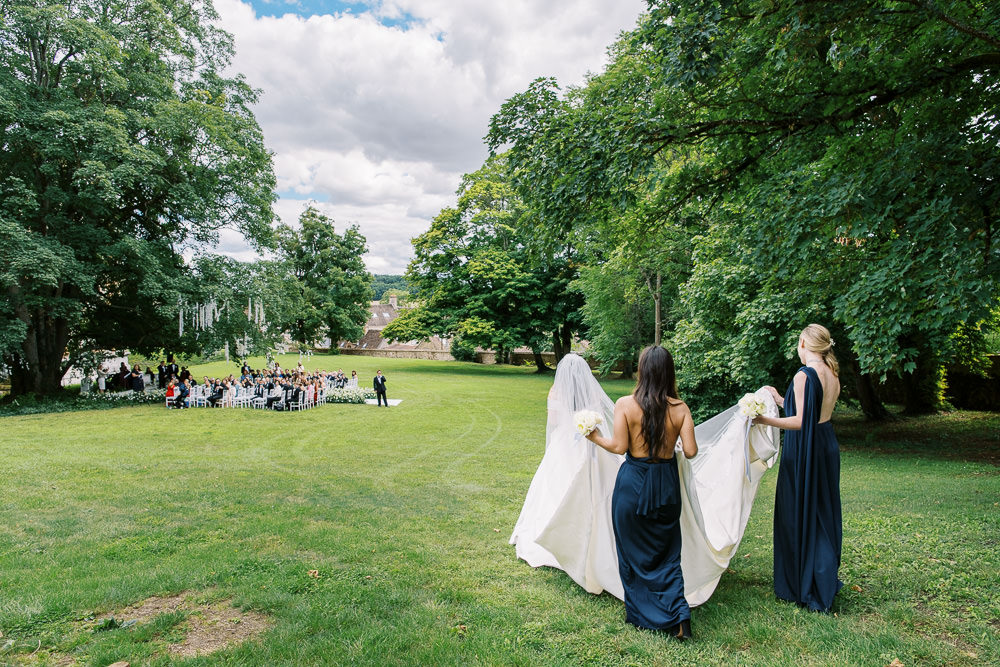 A bride in a white gown with a long cathedral veil and train is walking across a large open lawn toward an outdoor ceremony in the distance, accompanied by two bridesmaids in navy blue floor-length halter-style dresses who are carrying her train and holding white bouquets. The ceremony setup in the background includes rows of white chairs filled with approximately 100 guests, with the groom visible at the altar flanked by an officiant. The setting appears to be the grounds of a French chateau or estate, with historic stone buildings partially visible through the trees in the background. The wide-angle shot is taken from behind the bridal party, capturing the full expanse of the lawn and the procession in progress.