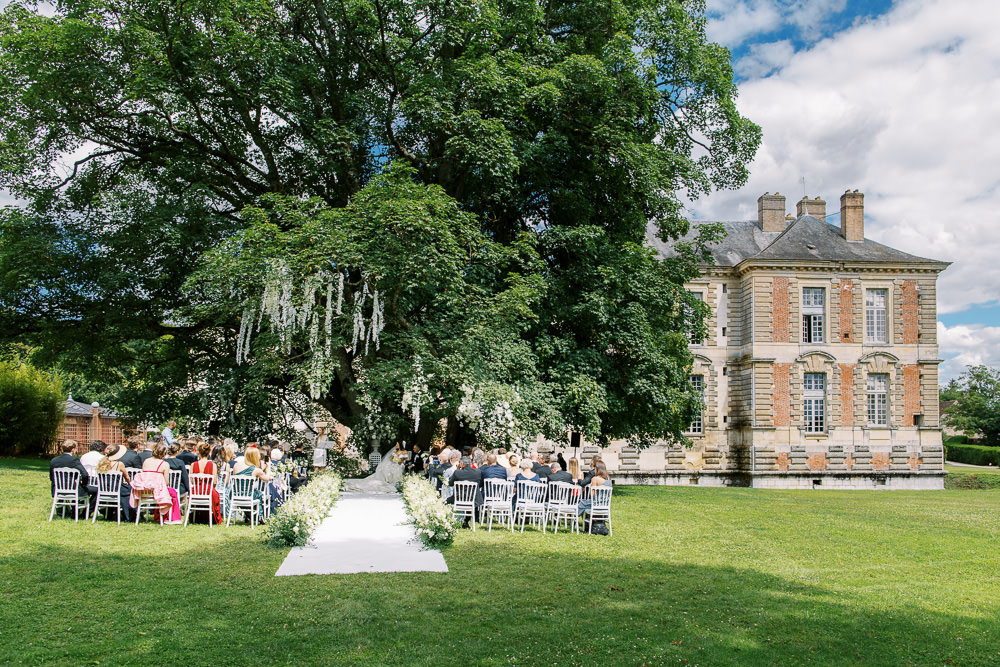 An outdoor wedding ceremony taking place on the grounds of a French chateau, with approximately 80–100 guests seated in white Chiavari chairs arranged in two sections facing a natural altar formed beneath a large mature tree. The white aisle runner is flanked by low arrangements of white flowers and greenery. White floral garlands or hanging installations cascade from the tree branches above the ceremony space, creating a canopy effect. The chateau, a classic French country house in stone and red brick with mansard roof and chimney stacks, is visible to the right of the frame. Wide-angle shot taken from behind the guests, capturing the full ceremony layout and the chateau facade in one composition. Potential venue feature image.