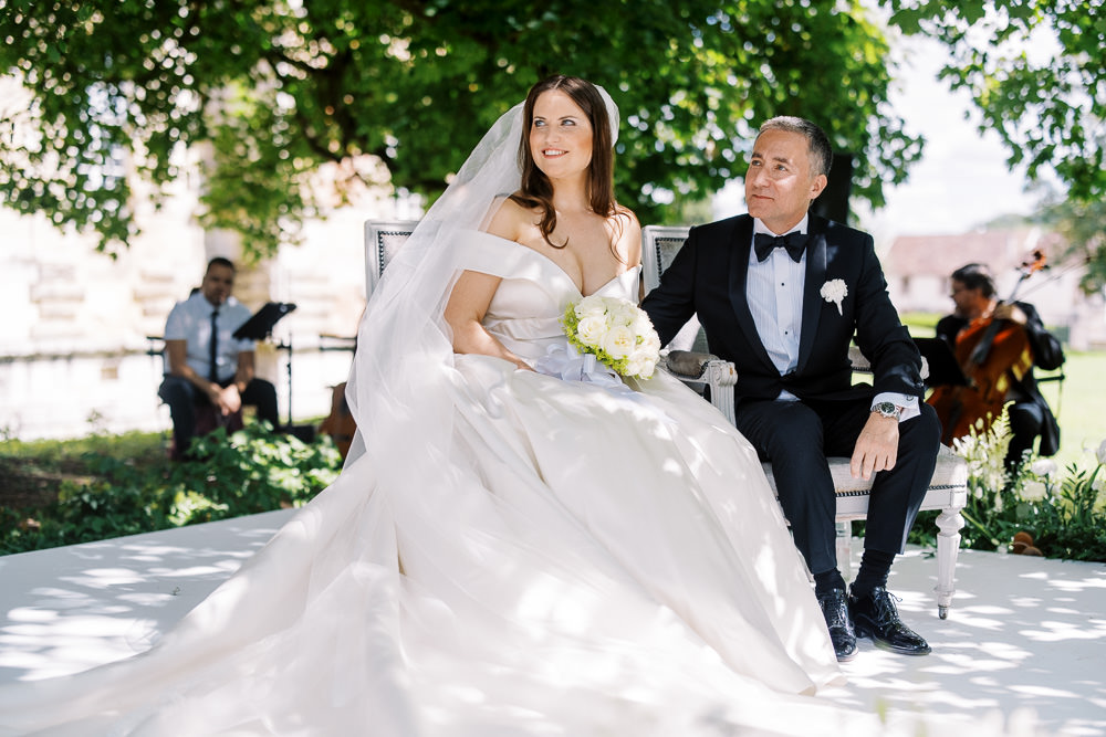The bride and groom are seated on a white French-style settee during an outdoor ceremony, set on a white platform stage under the shade of a large tree. The bride wears an off-the-shoulder ivory ballgown with a voluminous skirt and a long cathedral veil, holding a rounded bouquet of white roses and green accents. The groom is dressed in a classic black tuxedo with a bow tie and a white boutonnière. Two live musicians — one with sheet music and one playing cello — are visible in the background, suggesting a live string ensemble performing during the ceremony. The setting appears to be the grounds of a chateau or historic estate, with stone architecture faintly visible in the distance. The overall styling is classic and formal, with a white and ivory decor palette. The shot is a mid-range portrait capturing both the couple and the ambient ceremony scene.
