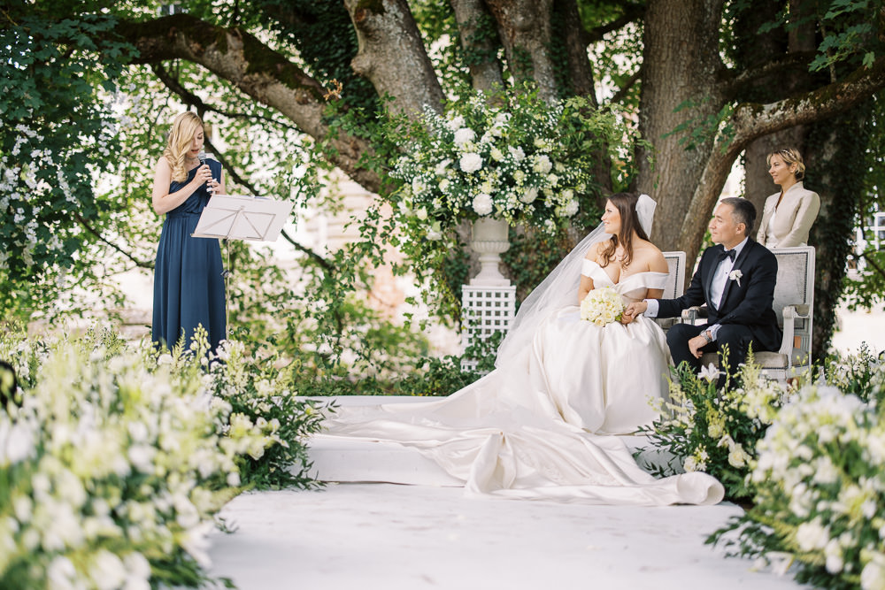 An outdoor wedding ceremony taking place beneath a large mature tree, with the couple seated in upholstered chairs on a raised white platform. The bride wears an off-the-shoulder full ballgown in ivory with a long cathedral-length veil and holds a bouquet of white hydrangeas and cream roses, while the groom is dressed in a black tuxedo with a white boutonniere. A woman in a floor-length navy blue dress reads from a music stand at a podium to the left, likely delivering a reading, while a female officiant in a cream blazer stands behind the couple. The ceremony altar features a white stone urn overflowing with white hydrangeas, garden roses, and greenery, and the aisle is lined on both sides with lush arrangements of white snapdragons, white blooms, and trailing greenery. The overall decor palette is white and ivory with deep green foliage, giving a classic, garden-style aesthetic, captured in a wide editorial shot framing the full scene.