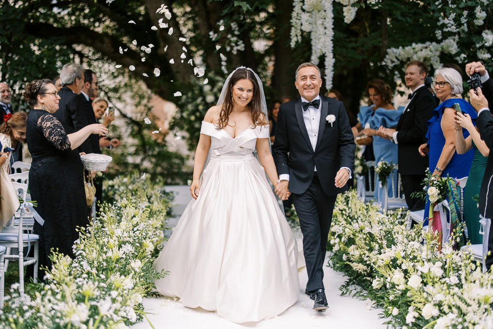The bride and groom walk back down the aisle together following an outdoor ceremony, with guests on both sides tossing white flower petals over them. The bride wears an off-the-shoulder ivory ballgown with a full satin skirt and a cathedral-length veil, while the groom is dressed in a black tuxedo with a bow tie and a white boutonnière. The aisle is lined with lush arrangements of white flowers — including what appear to be white lisianthus, freesia, and greenery — laid directly on the ground, and a floral arch with hanging white blooms is visible in the background. The ceremony took place in a garden or chateau grounds setting, and approximately 20–30 guests are visible, some in formal attire including a woman in a cobalt blue dress; the mid-length shot captures the couple centered and smiling, with natural daylight illuminating the scene.