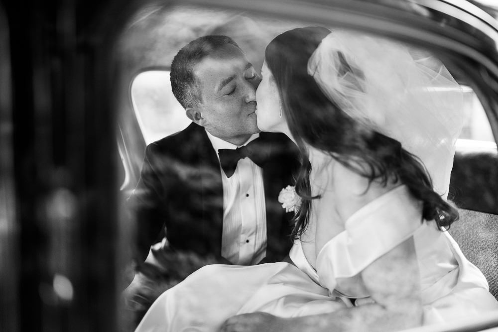 A black-and-white portrait of a bride and groom kissing in the back seat of a vintage car. The groom wears a classic tuxedo with a bow tie and a small floral boutonnière on his lapel, while the bride wears a white satin gown and a long veil that billows across the frame. The image is shot through the car window, creating soft reflections and lens flare effects that partially overlay the couple. The composition is a close-up portrait with high contrast black-and-white tones, and the veil movement adds a sense of texture and depth to the frame.