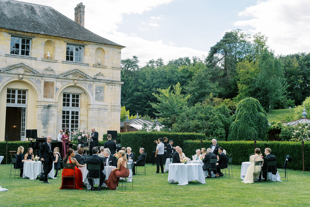 An outdoor cocktail hour or garden reception taking place on a manicured lawn beside a classical French chateau with pale yellow stone facade, arched windows, and ornamental architectural detailing. Approximately 30–40 guests are seated and mingling at round tables dressed in white linens, with black metal chairs; a small floral arrangement in blush and coral tones is visible on one table. Guests are dressed in formal attire including black tie suits, a deep red/burgundy gown, a burnt orange dress, and a champagne-colored dress. A grand piano and what appears to be a small musical ensemble are set up near the building's exterior wall, with a tall floral arrangement in blush and white tones alongside. Wide shot capturing both the venue architecture and the full garden scene. Potential venue feature image.