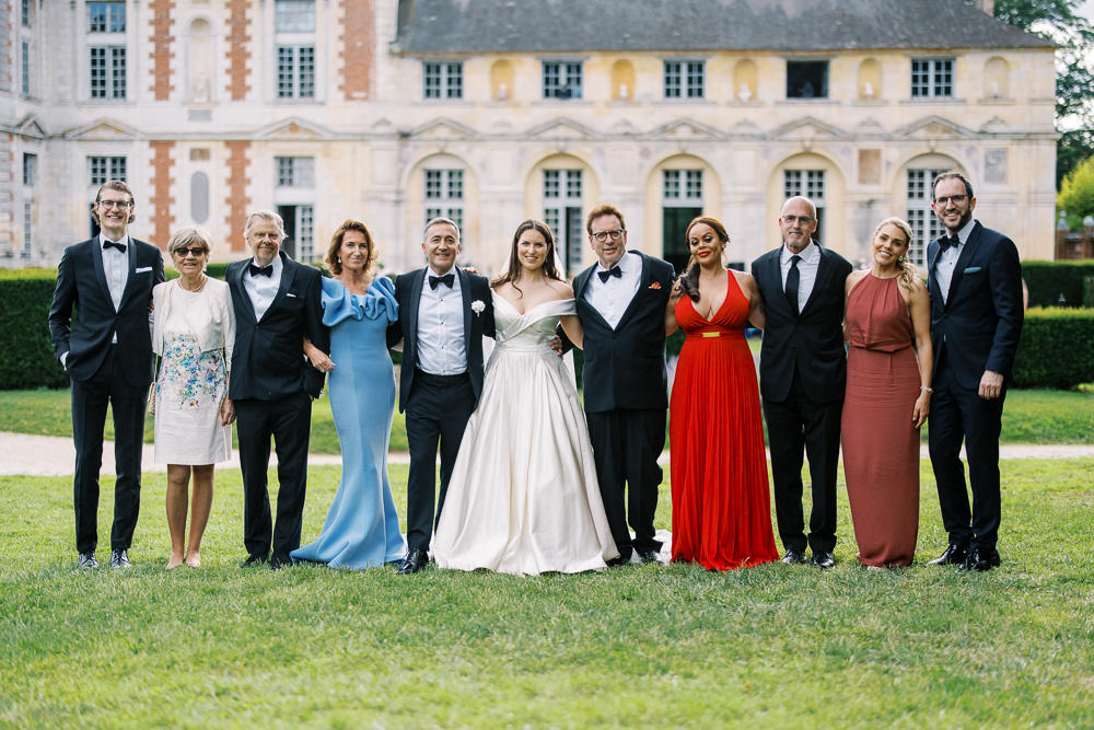 A group portrait of eleven people posing outdoors on the grounds of a French chateau, with the classic brick-and-stone facade of the building visible in the background. The bride stands at the center wearing an off-the-shoulder ivory satin ball gown, and the groom beside her is dressed in a black tuxedo with a white boutonniere. The group includes men in black tuxedos and women in a variety of formal dresses: a light blue gown with ruffled shoulders, a red deep-V halter gown with a gold waist accent, a dusty mauve halter-neck gown, a short white floral-print dress, and a navy dress partially visible. The shot is a wide, evenly-lit daytime group portrait with all subjects standing in a single line facing the camera. Potential venue feature image.