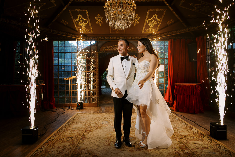 The couple stands together on an ornate patterned rug during what appears to be a reception entrance or first dance moment, framed by two cold sparkler fountains on either side. The groom wears a white dinner jacket with black bow tie and black trousers, while the bride wears a strapless hi-low white gown with beaded embellishments on the bodice and layered organza skirt. The indoor venue features a dark ceiling with gold ornamental molding, a crystal chandelier, large arched French windows, and deep red draped curtains to the right, suggesting a classic, formal ballroom setting. The composition is a wide portrait shot with the sparklers creating dramatic framing light against the dark room.