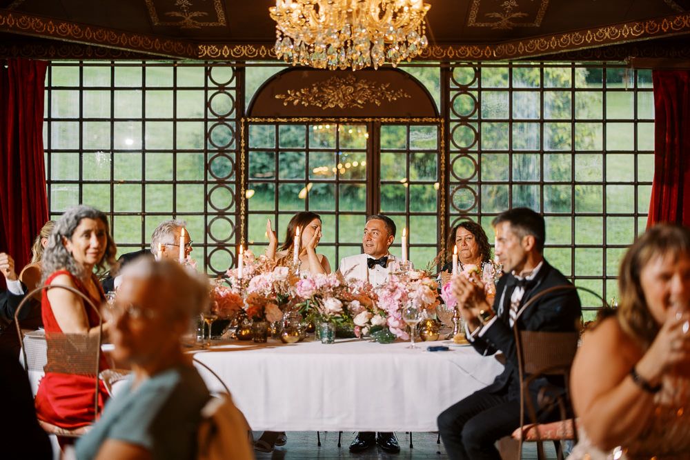 A wedding reception dinner is underway inside a formal ballroom or chateau dining room, with the couple seated at a sweetheart table during what appears to be speeches. The groom wears a white tuxedo jacket with black bow tie, and the bride appears to be covering her mouth in an emotional or laughing reaction. The room features ornate gold-detailed dark wood paneled windows, rich red velvet drapes, a crystal chandelier overhead, and a decorative gold-painted ceiling. The sweetheart table is dressed with a white linen and an abundant low floral arrangement in shades of blush pink, coral, and peach — likely including garden roses and ranunculus — with white taper candles and gold accent vessels. Approximately eight to ten guests are visible seated at surrounding tables, with one woman in a red dress prominently in the foreground. Wide shot capturing the full scene with the couple centered against the large window backdrop.