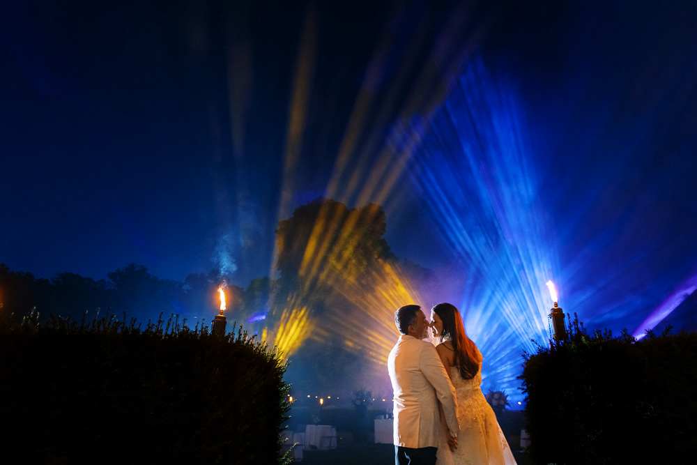 A nighttime outdoor couple portrait showing the bride and groom facing each other in an intimate near-kiss pose, backlit against a dramatic professional light show featuring radiating beams in gold, blue, and purple shooting upward into the dark sky, with atmospheric haze enhancing the light rays. The groom wears a white suit and the bride wears a white strapless embellished gown with long auburn hair. Two lit torch flares are visible flanking the couple on either side, positioned along trimmed hedgerows, with faint event lighting visible in the background suggesting a reception setting. The composition is a wide environmental portrait that places the couple as silhouetted figures against the large-scale lighting display, creating strong contrast between the dark foreground and the illuminated sky.