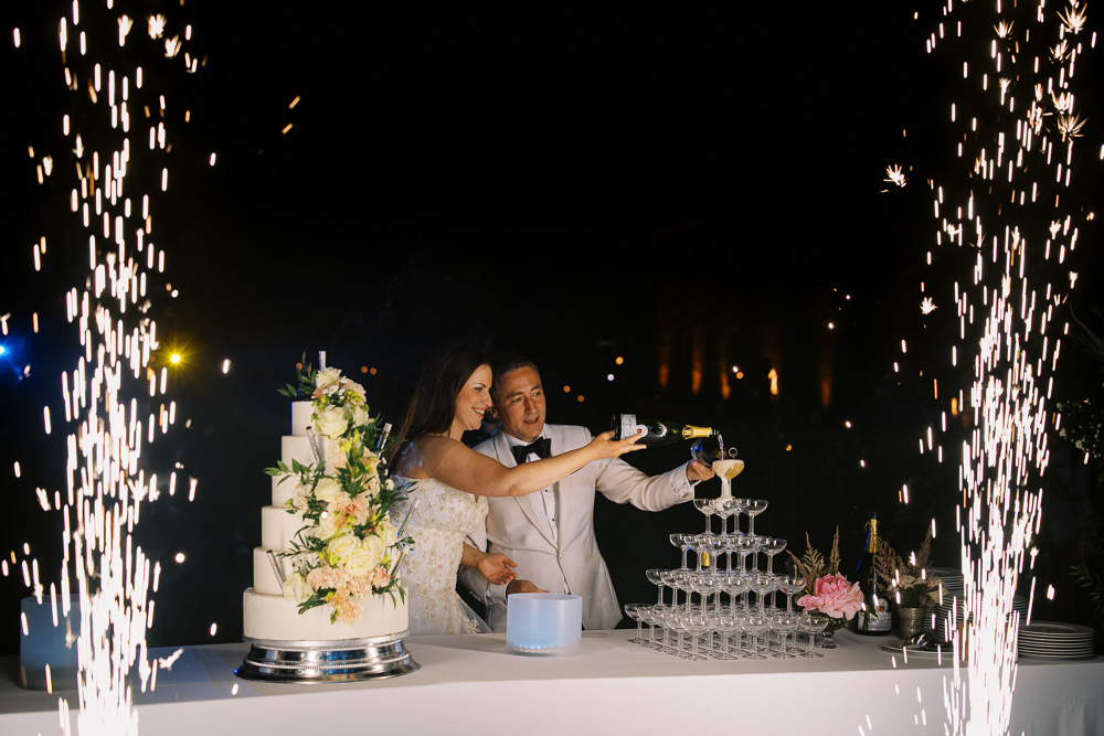 The couple stands at a white-linen table during an outdoor evening reception, pouring champagne into the top of a multi-tiered coupe glass tower while pyrotechnic cold sparkler fountains erupt on both sides of them. The bride wears an off-the-shoulder lace white gown and the groom wears a white dinner jacket with a black bow tie. To their left is a tall five-tier white wedding cake decorated with blush peach and ivory florals including roses and greenery, displayed on a silver cake stand. The table also holds a pink peony floral arrangement, stacked plates, and candles. The shot is a wide, slightly dark nighttime exposure with the sparklers creating dramatic streaks of light against the black background.